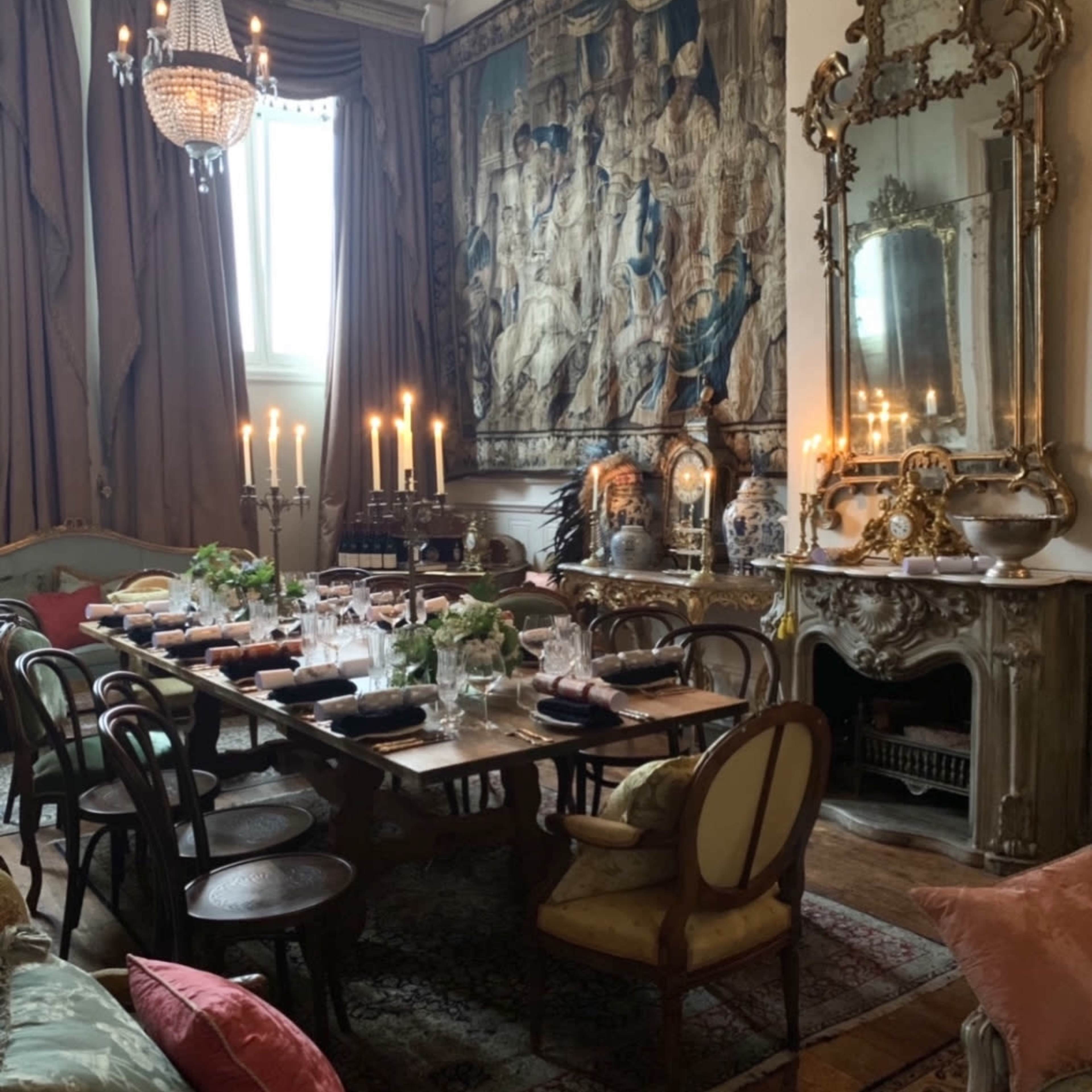 The picture shows an elegantly arranged dining room featuring a large wooden table set with dishes and candles, surrounded by various seating and decorated with a tapestry and a mirror.