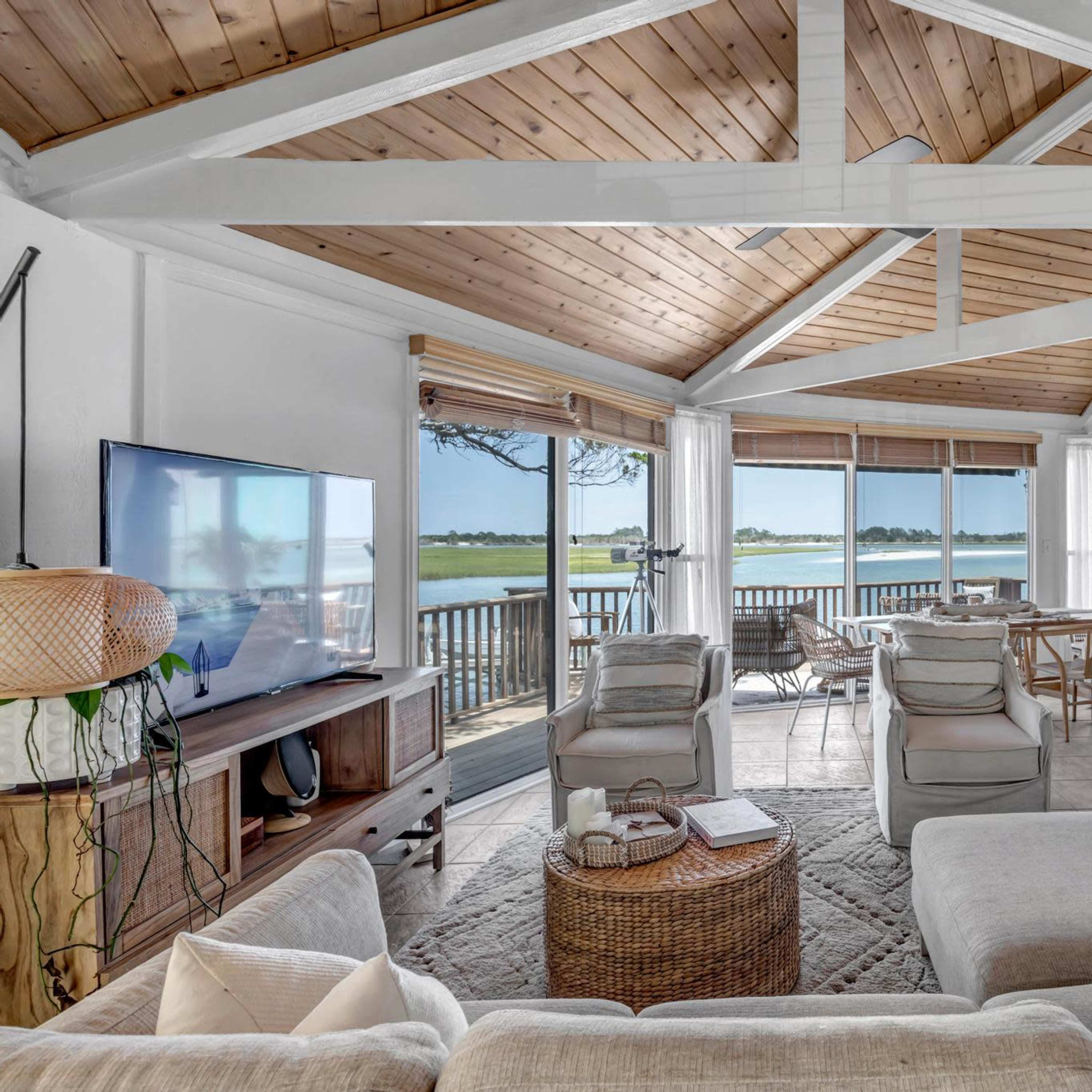 A spacious living room features a large window with a view of the water, complemented by wooden beams and a modern fireplace.