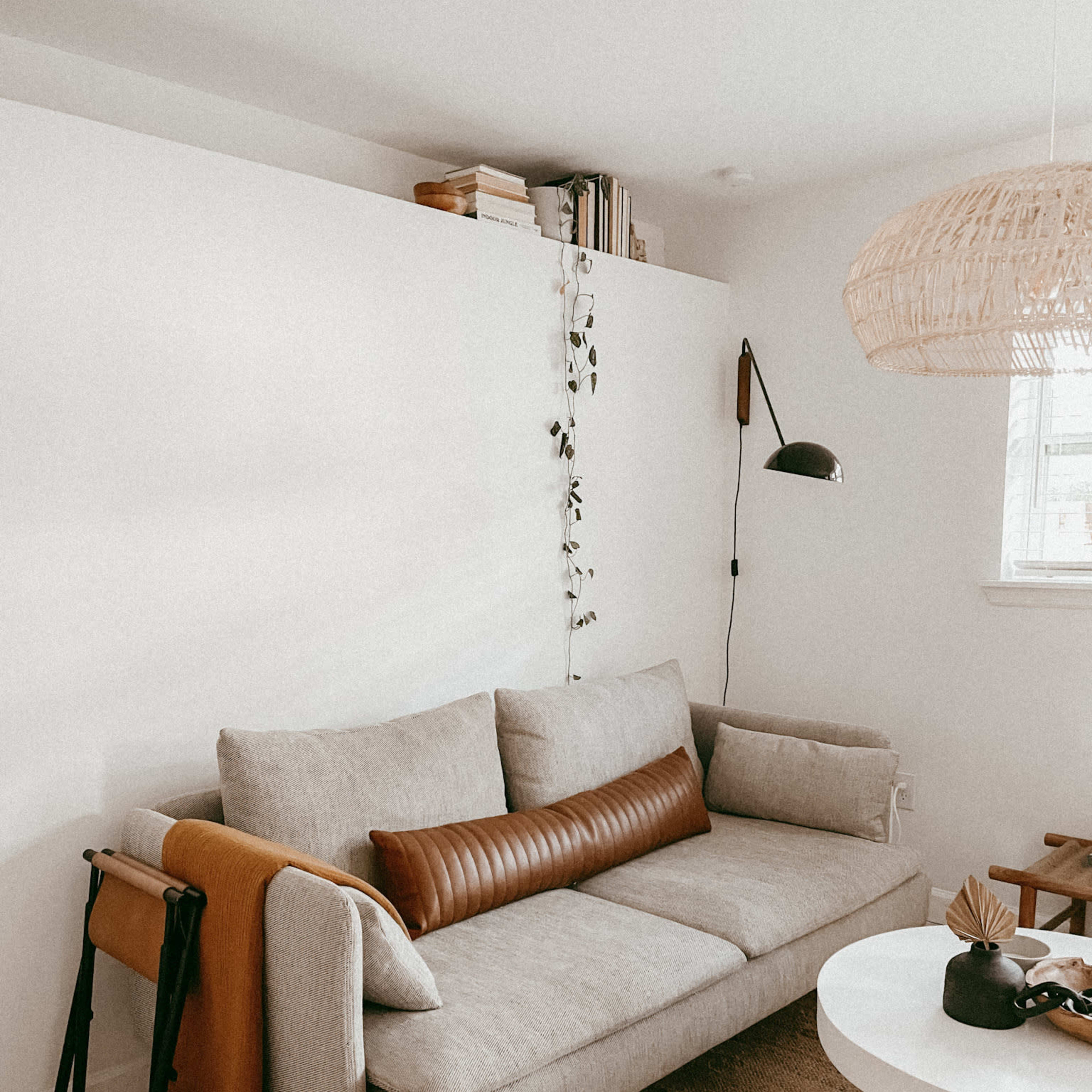 A light-colored living room features a gray sofa with a brown leather pillow, a round white coffee table, and a woven pendant light hanging above.