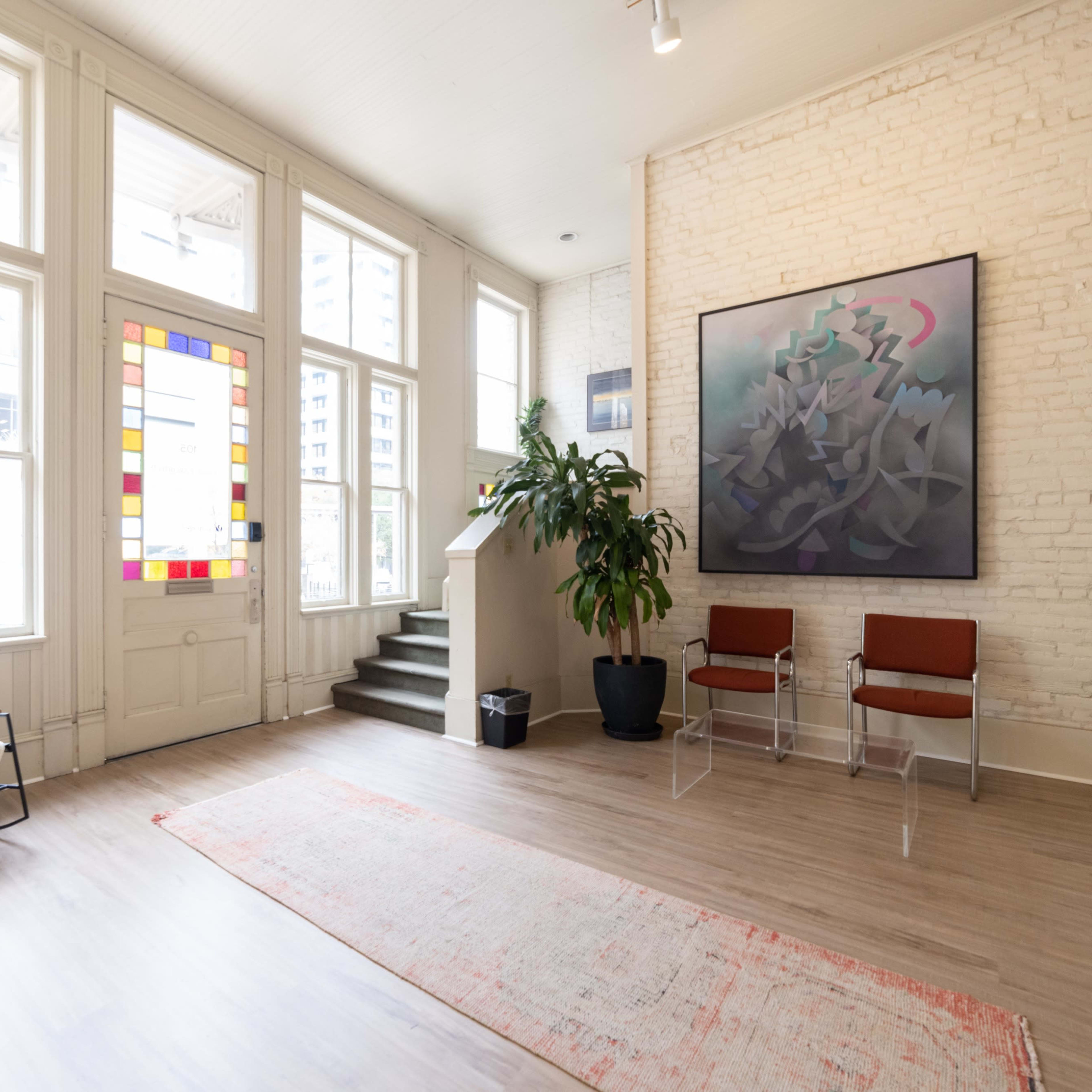 A spacious room with large windows, a colorful stained glass panel, a potted plant, two transparent chairs, and a patterned rug on a wooden floor.