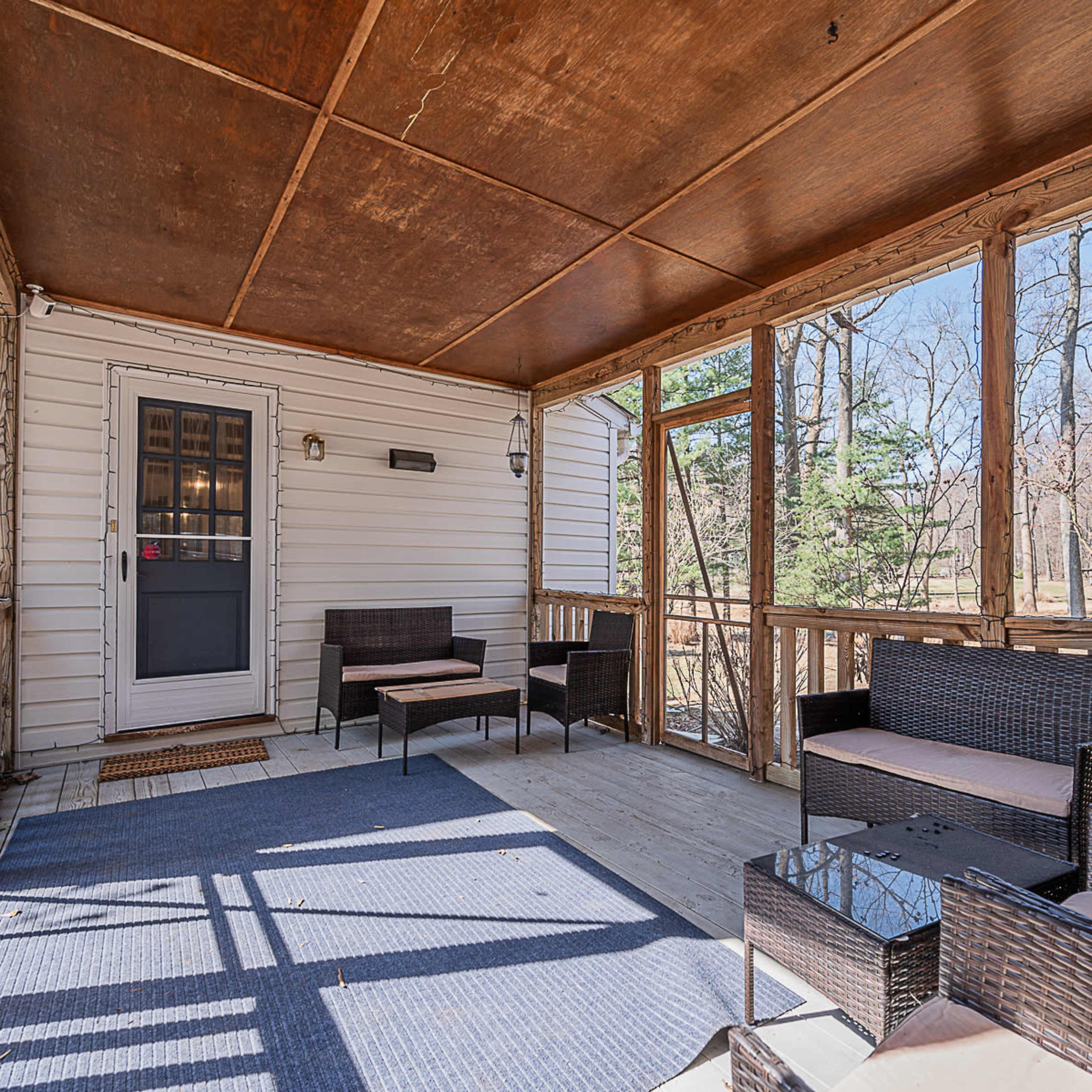 A screened porch with wicker furniture and a door leading outside, surrounded by trees in the background.