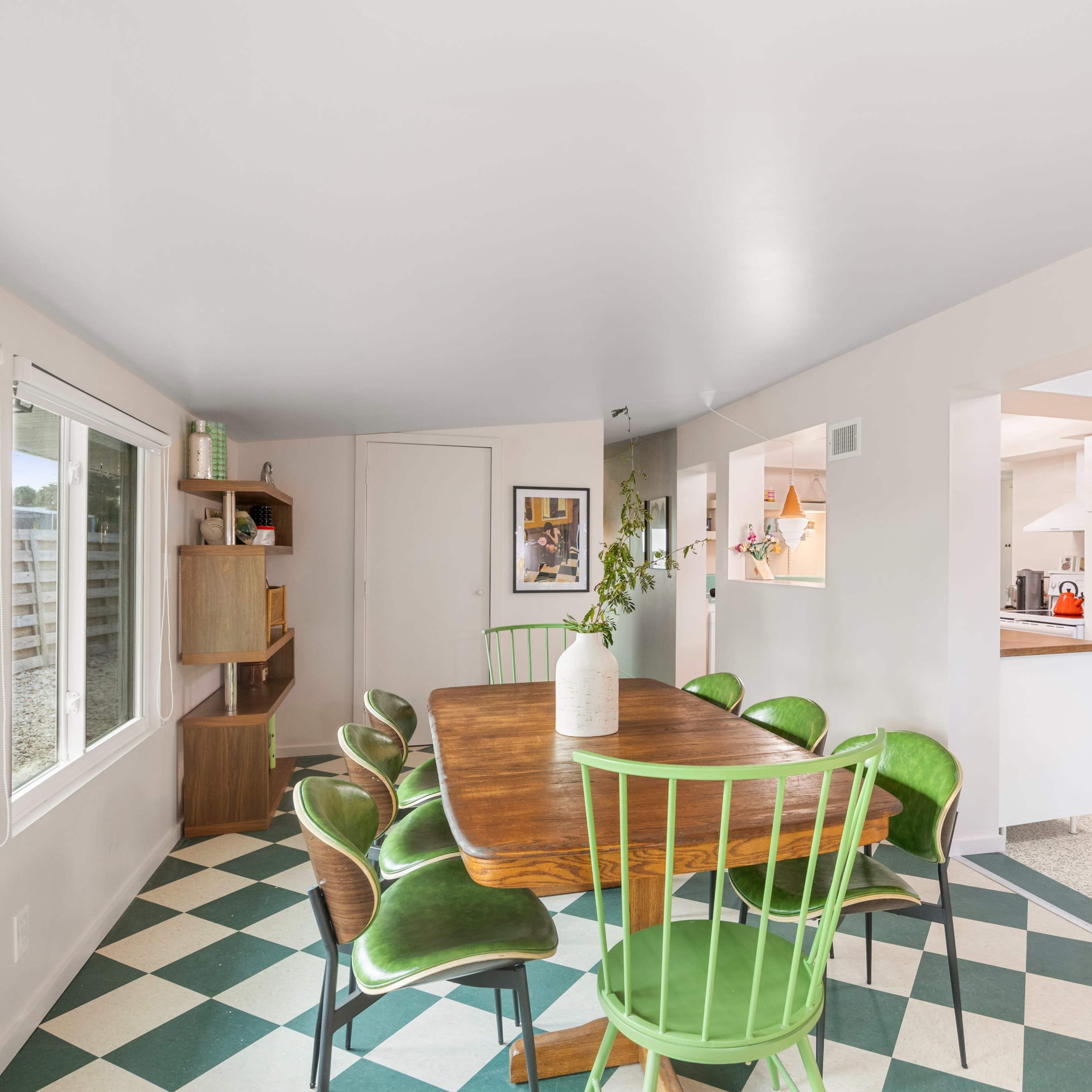 A dining area features a wooden table surrounded by green chairs, with large windows providing natural light and a glimpse into an adjoining kitchen.