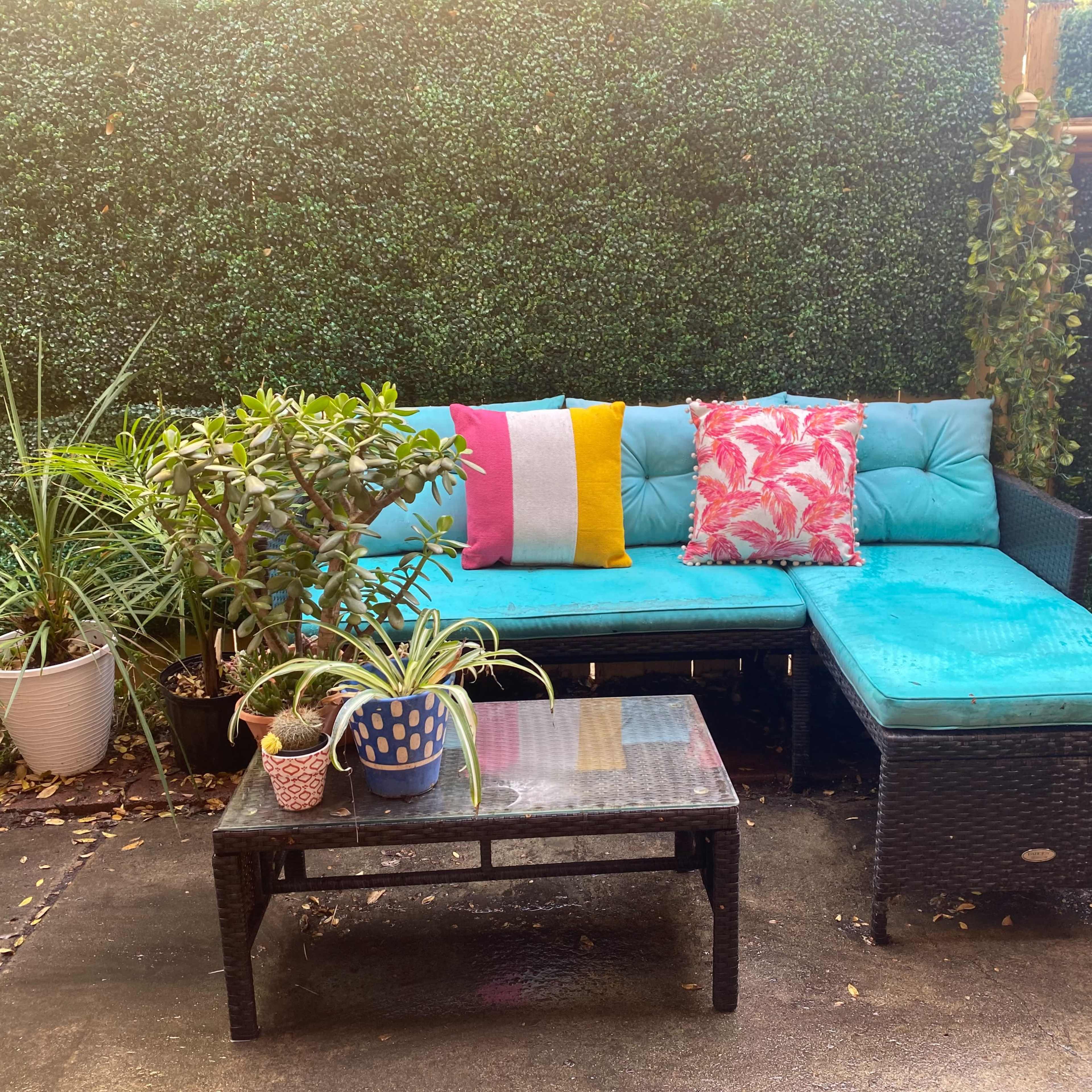 A seating area with a turquoise couch adorned with colorful cushions is surrounded by potted plants and a black coffee table on a concrete floor.