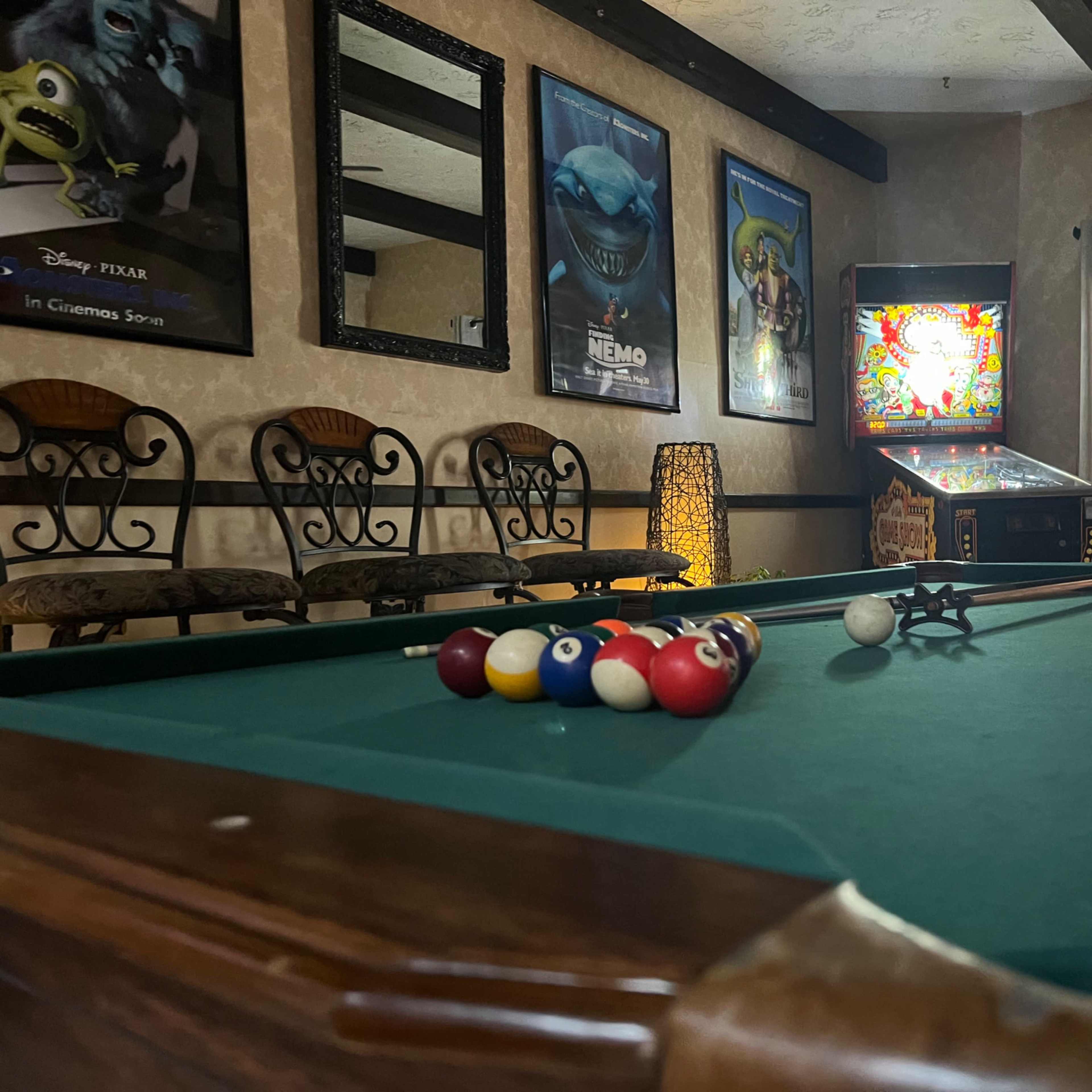 A billiards table is set up with balls racked at one end in a room featuring movie posters and an arcade machine.