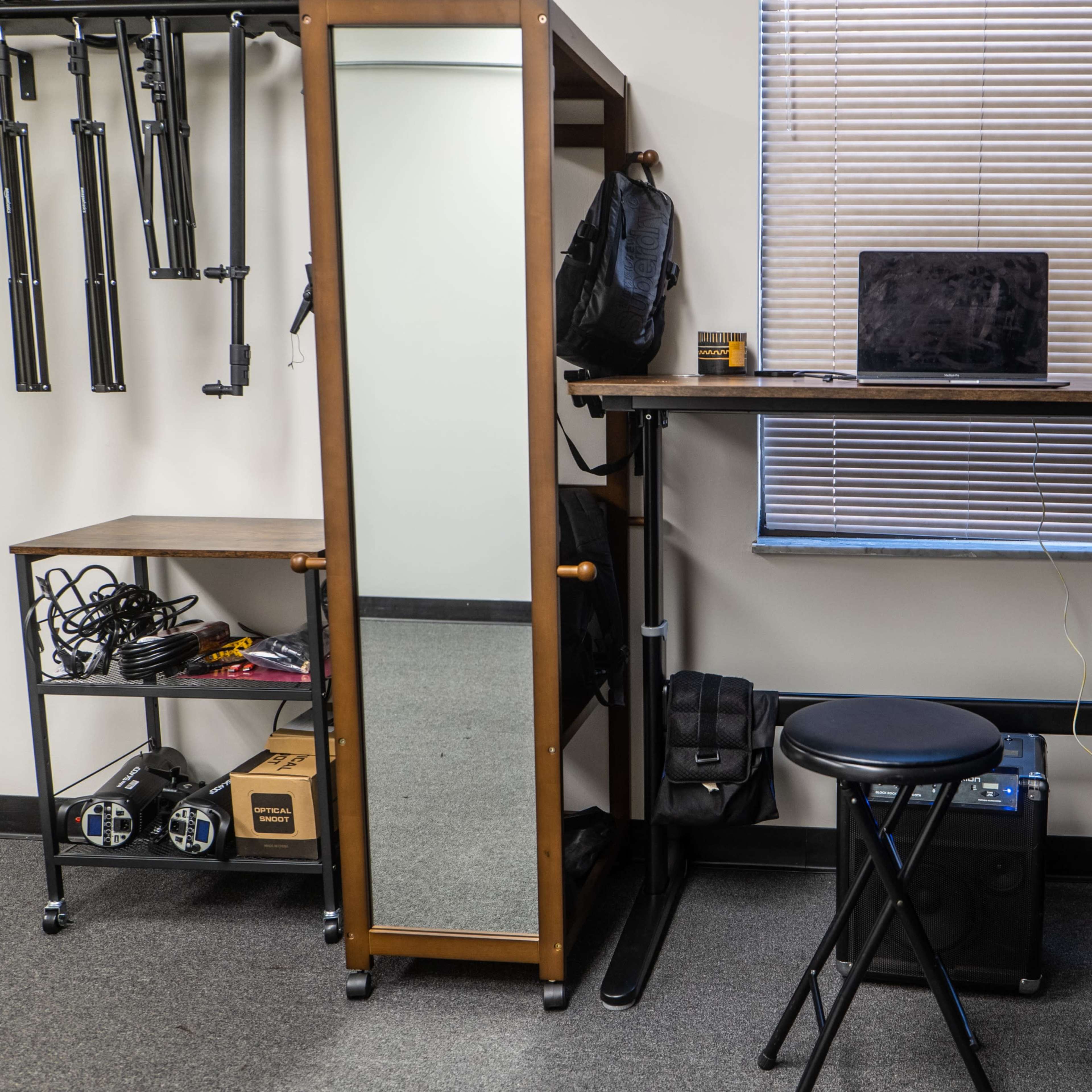A standing desk beside a mirror, a laptop on the desk, and a stool in front of it, with storage units and equipment visible in the background.