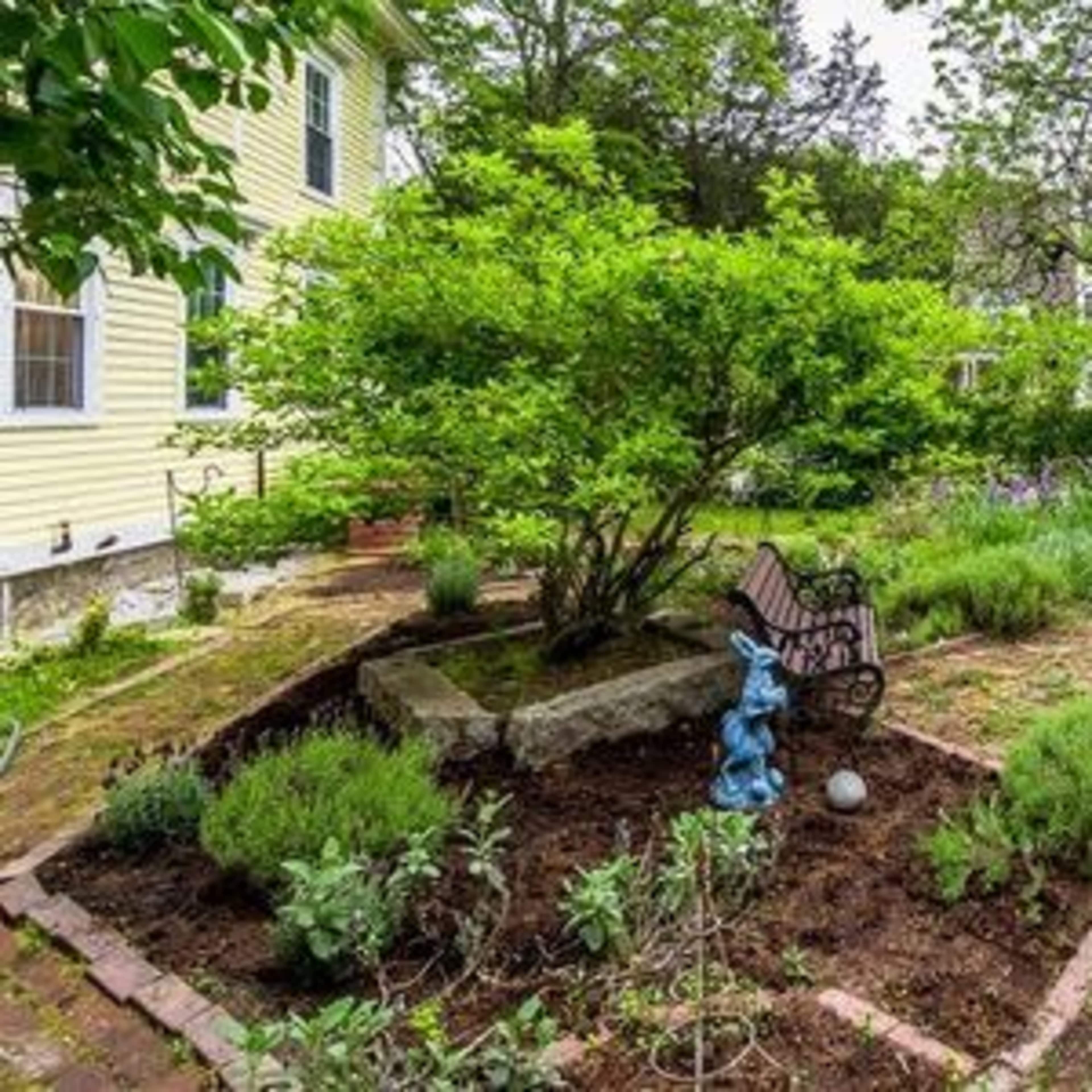 A garden features a green shrub at its center, surrounded by flower beds and a stone bench, with a blue rabbit statue nearby.