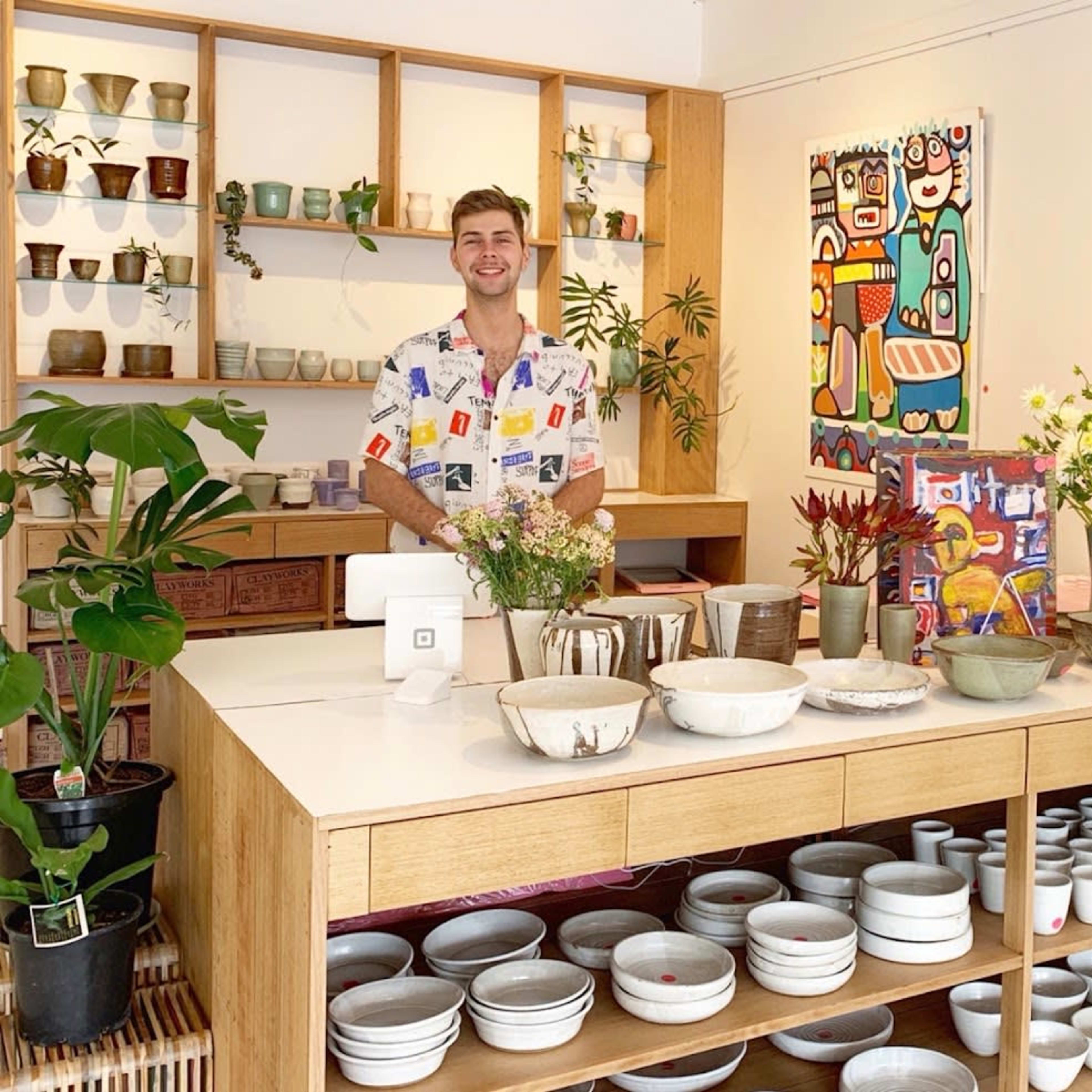 A bright gallery-style store features a smiling man standing behind a counter filled with various pottery and plants, while colorful artwork adorns the walls.