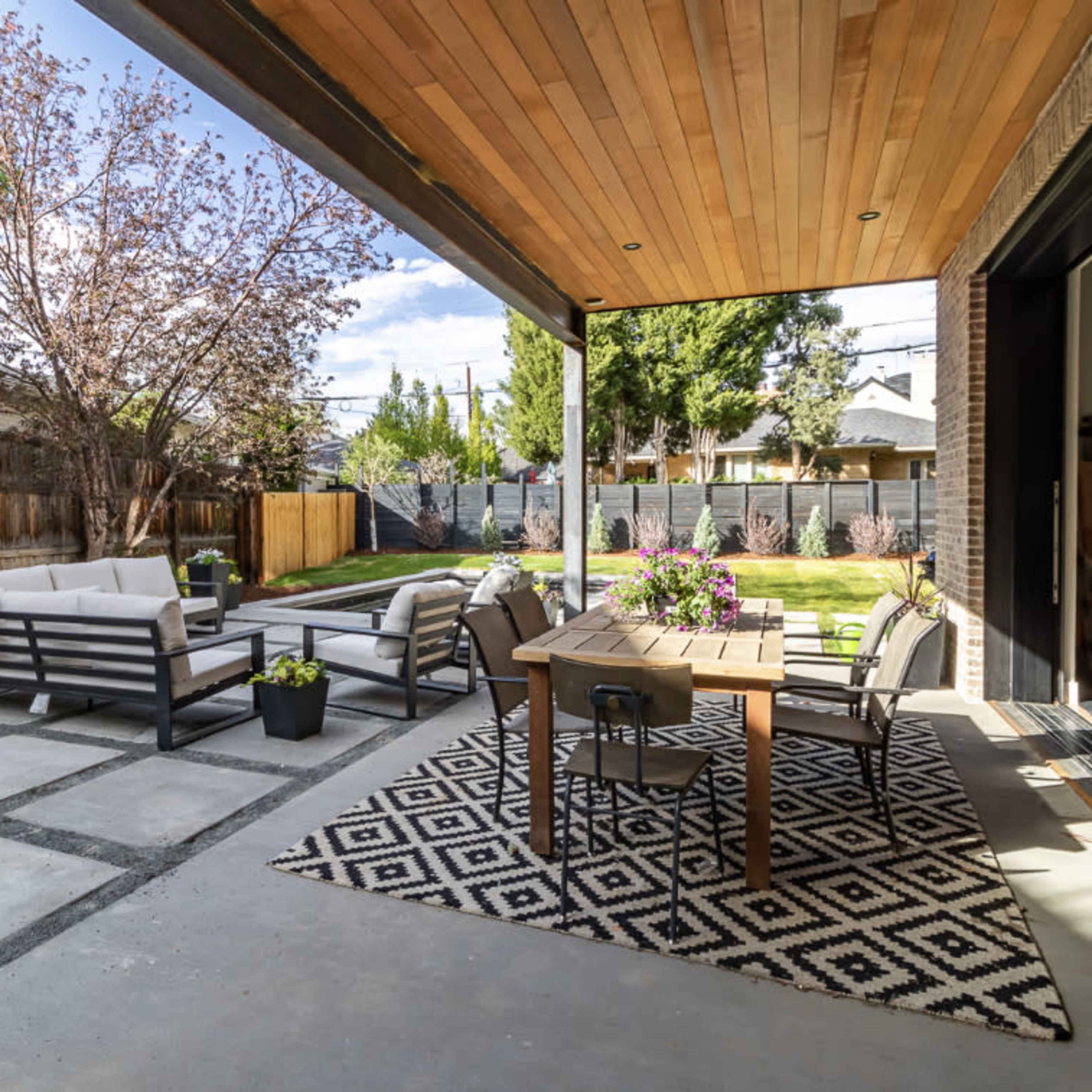 A patio area with outdoor seating, a dining table, and a garden space featuring trees and a lawn.
