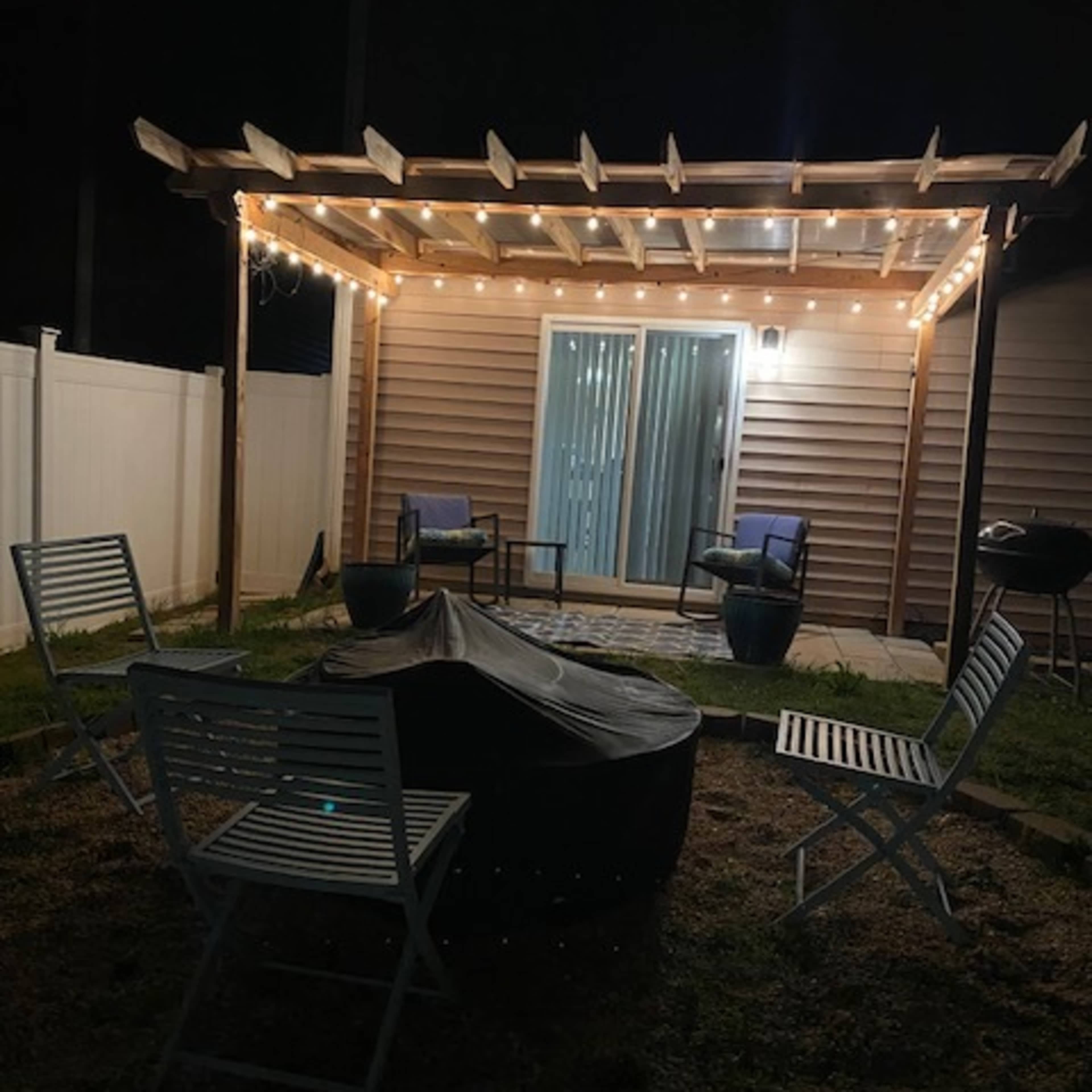 A backyard seating area features a pergola with string lights, surrounding a fire pit and several chairs.