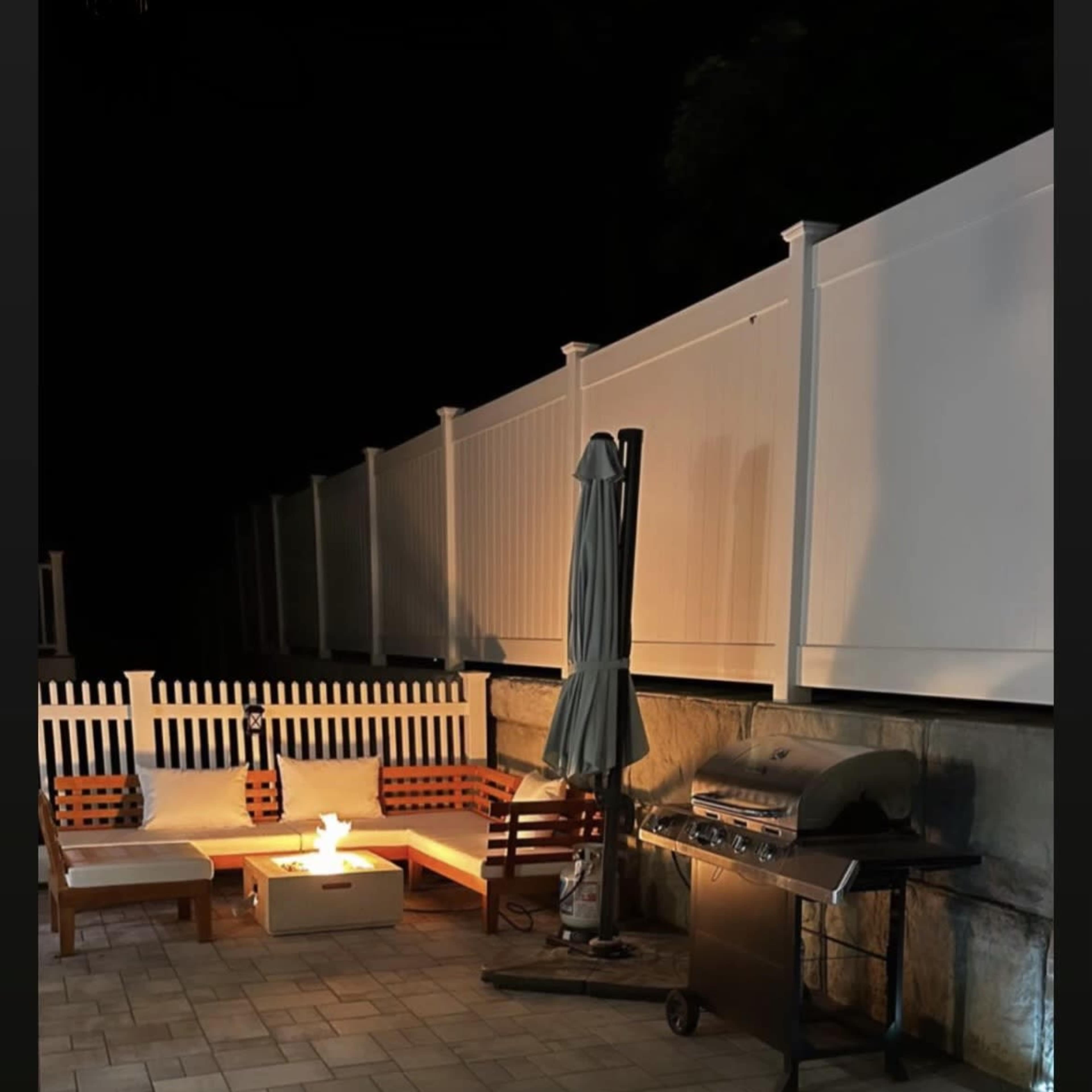 A patio area at night, featuring a fire pit surrounded by seating, a grill, and illuminated landscaping.