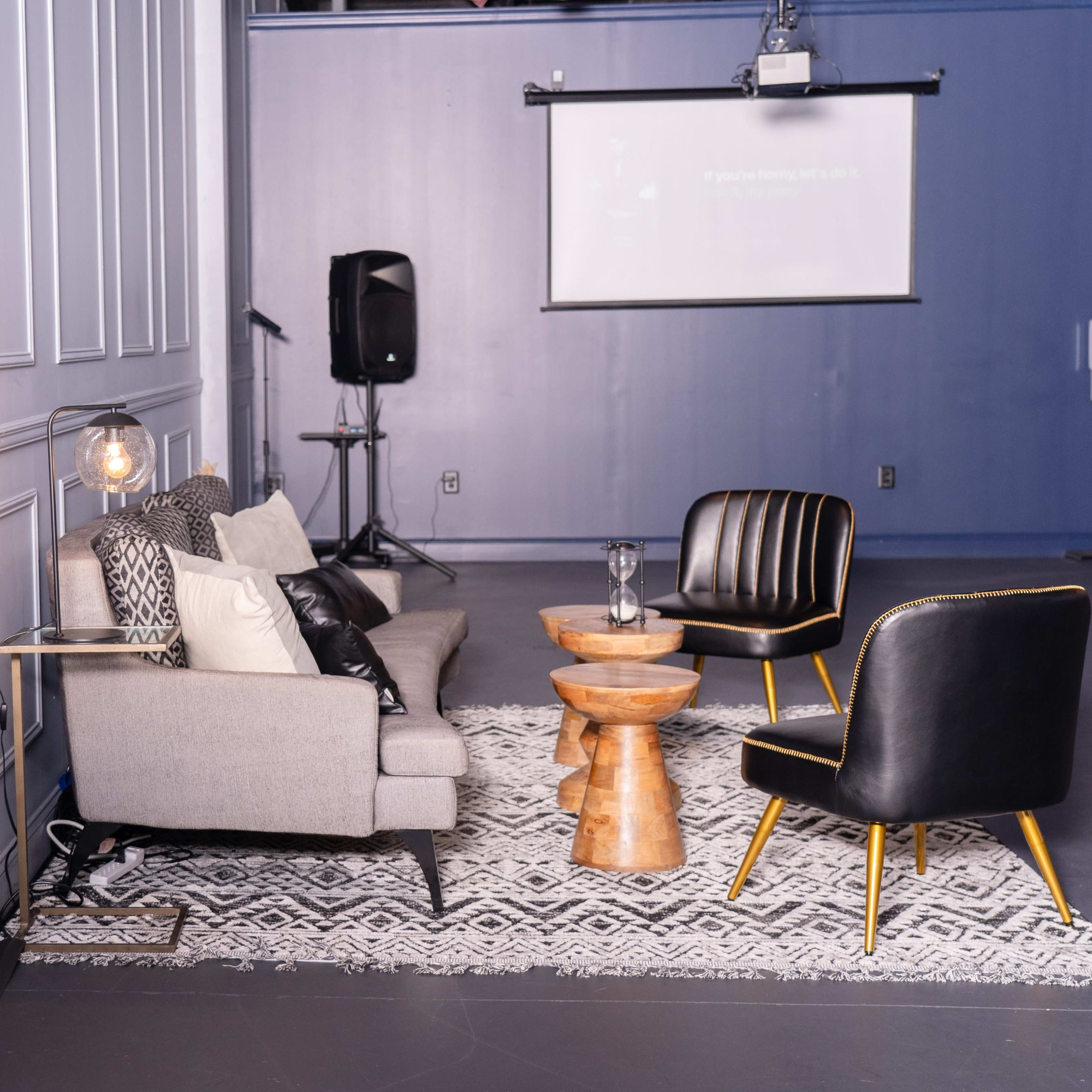 A stylish lounge area features a sofa, two black chairs, a wooden side table, and a projector screen in a modern space.