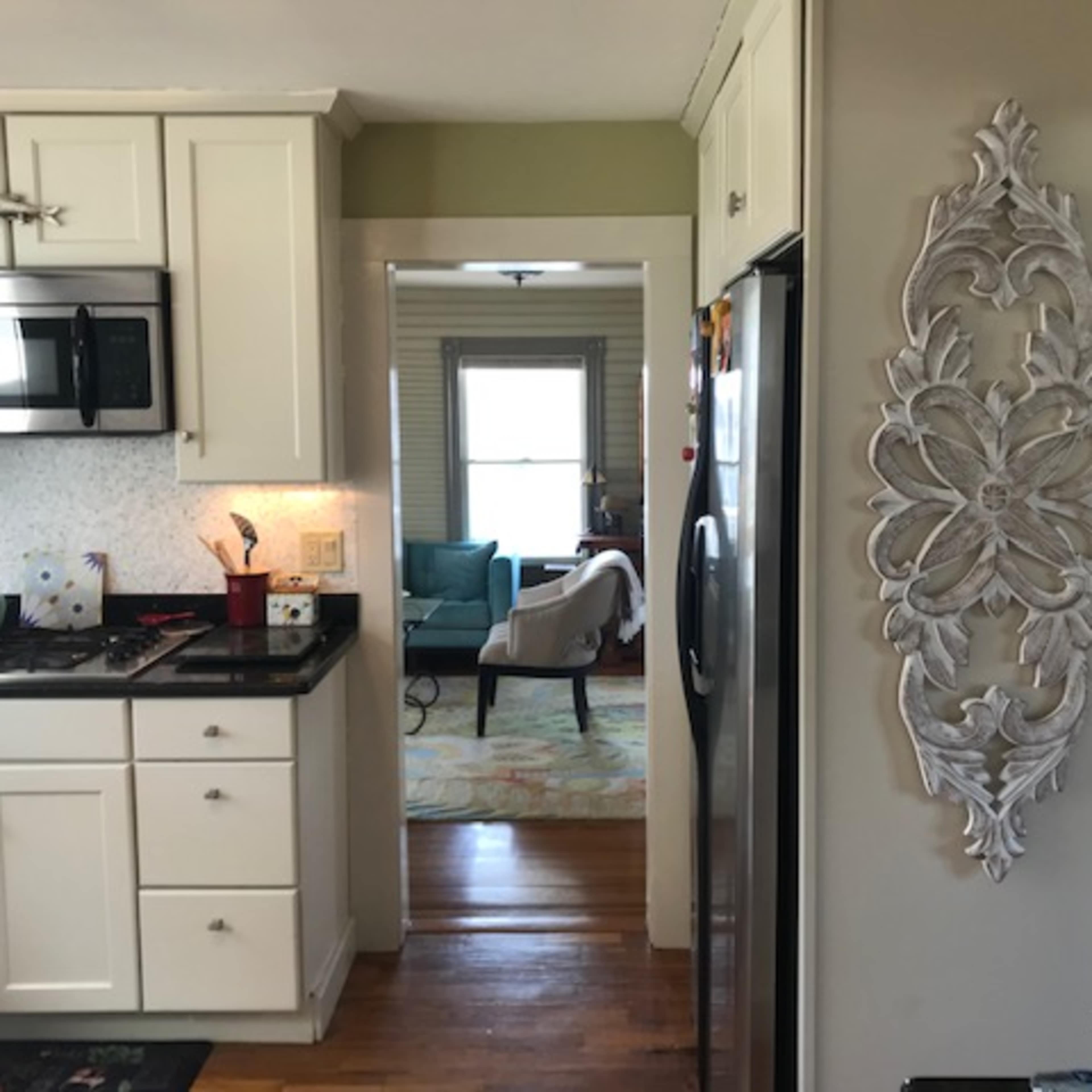 A kitchen with white cabinets and a stainless steel refrigerator, leading to a living area visible through an open doorway.