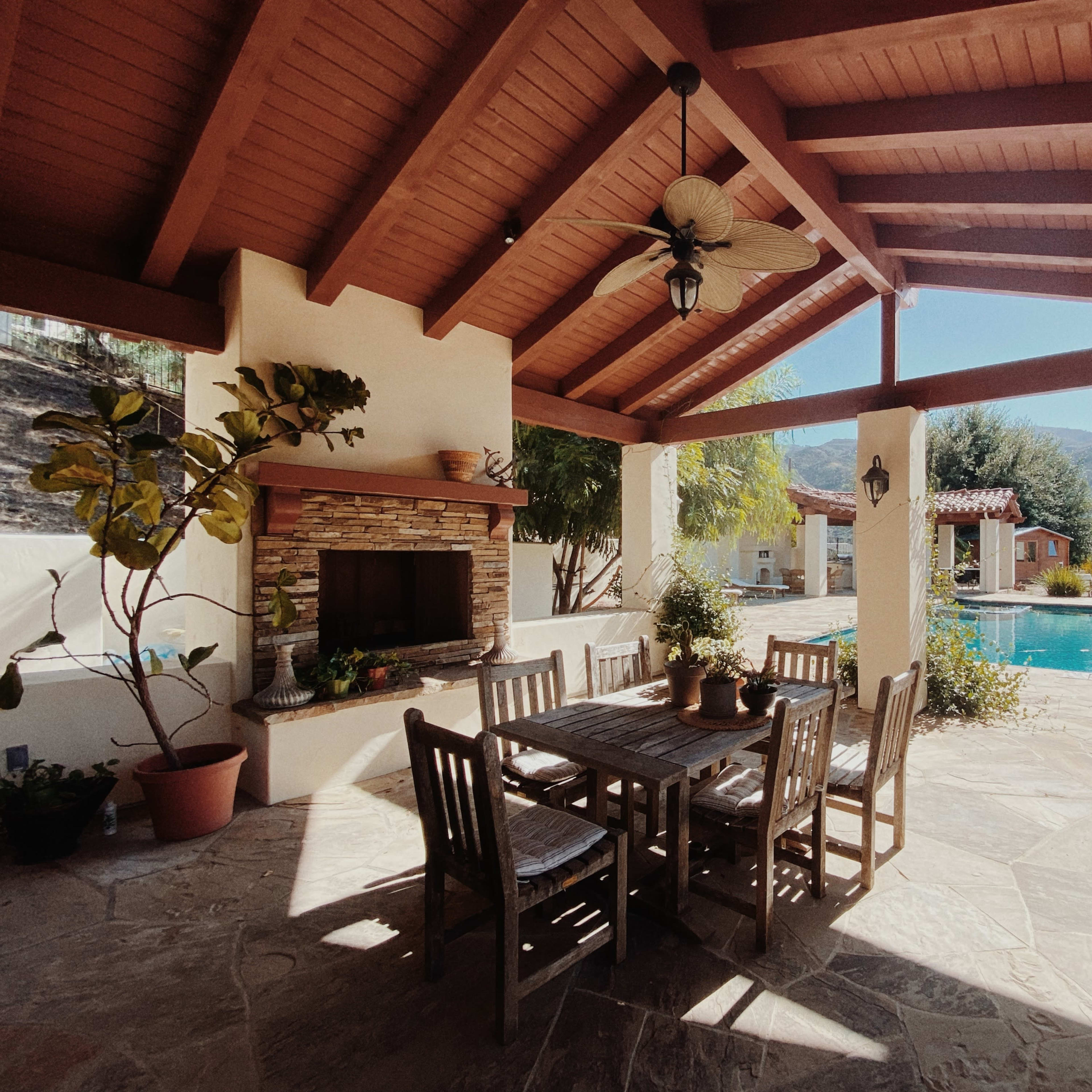 A covered patio features a wooden dining table surrounded by chairs, with a stone fireplace and a view of a swimming pool and mountains in the background.