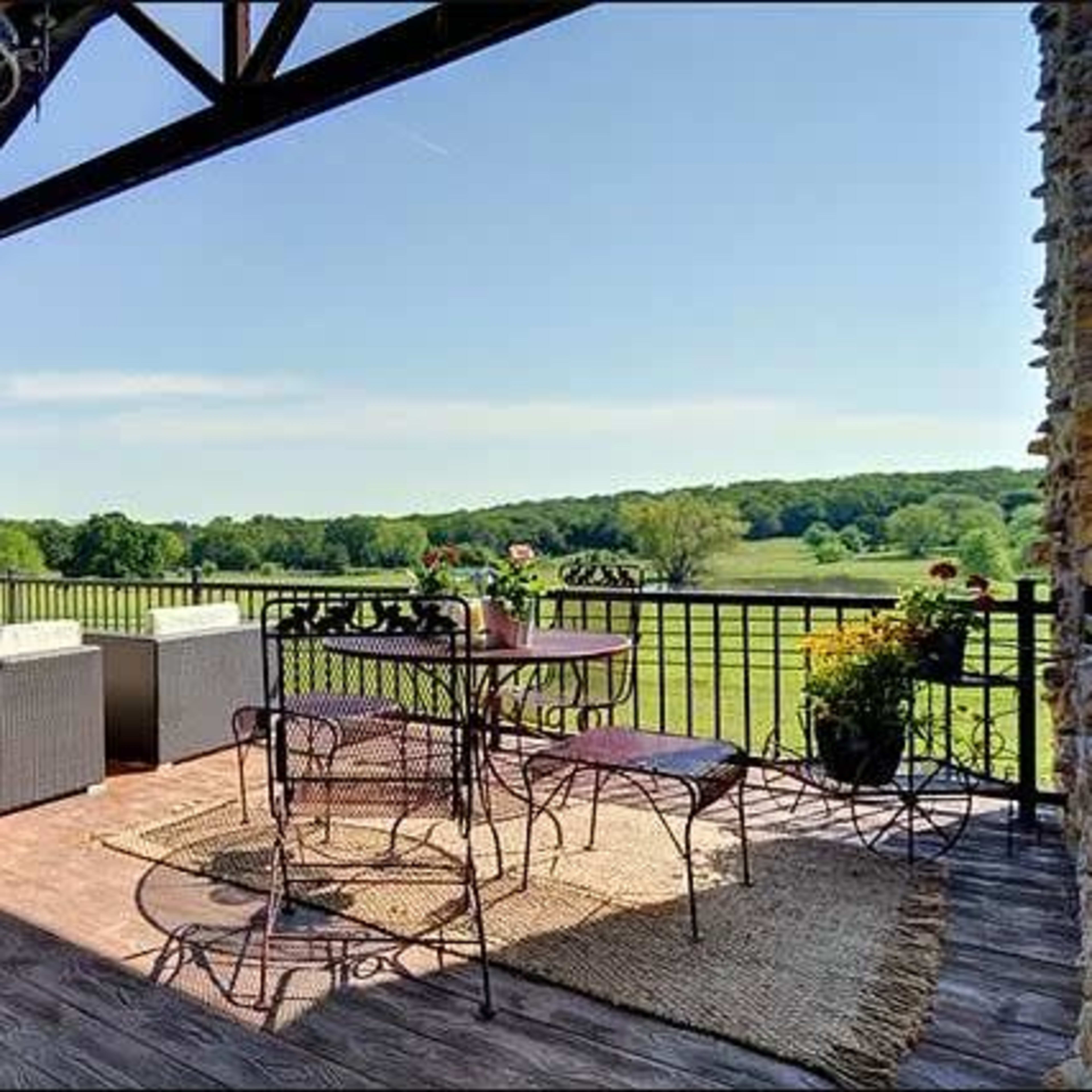 A covered patio features a seating area with wicker furniture and a dining table, overlooking a lush green landscape.