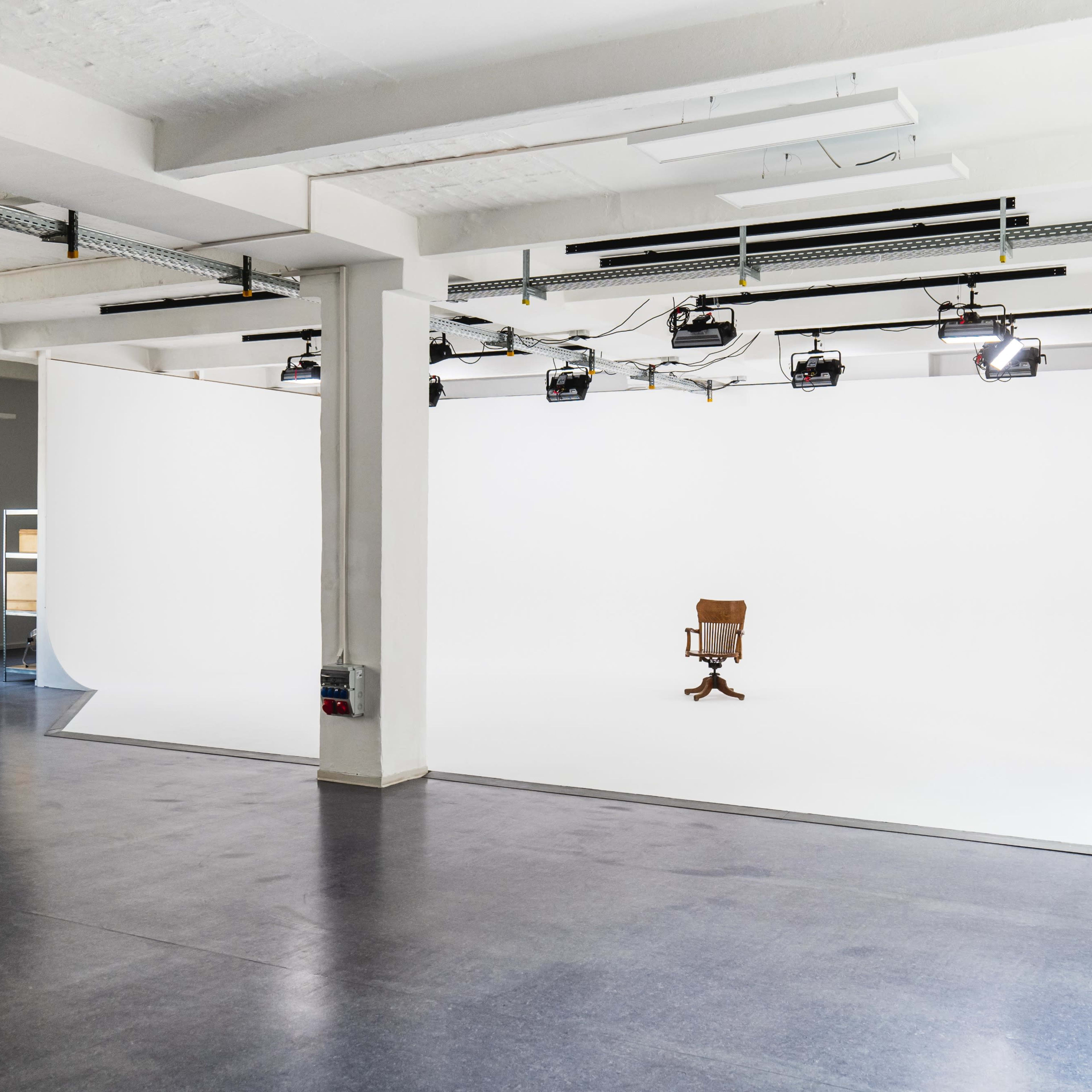 A spacious studio with a white backdrop, spotlights, and a single wooden chair positioned in the center.