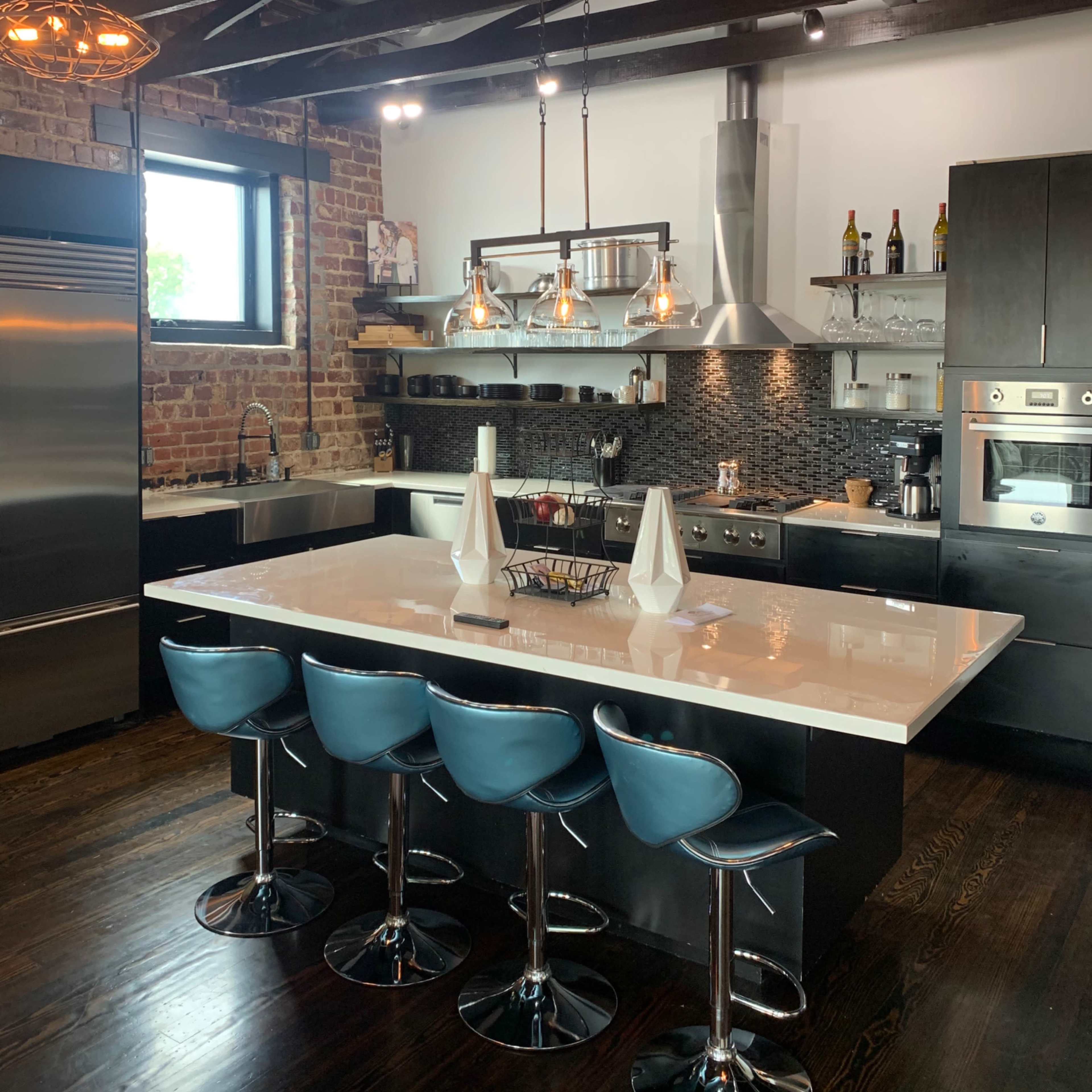 A modern kitchen features a large island with blue bar stools, exposed brick walls, pendant lighting, and stainless steel appliances.