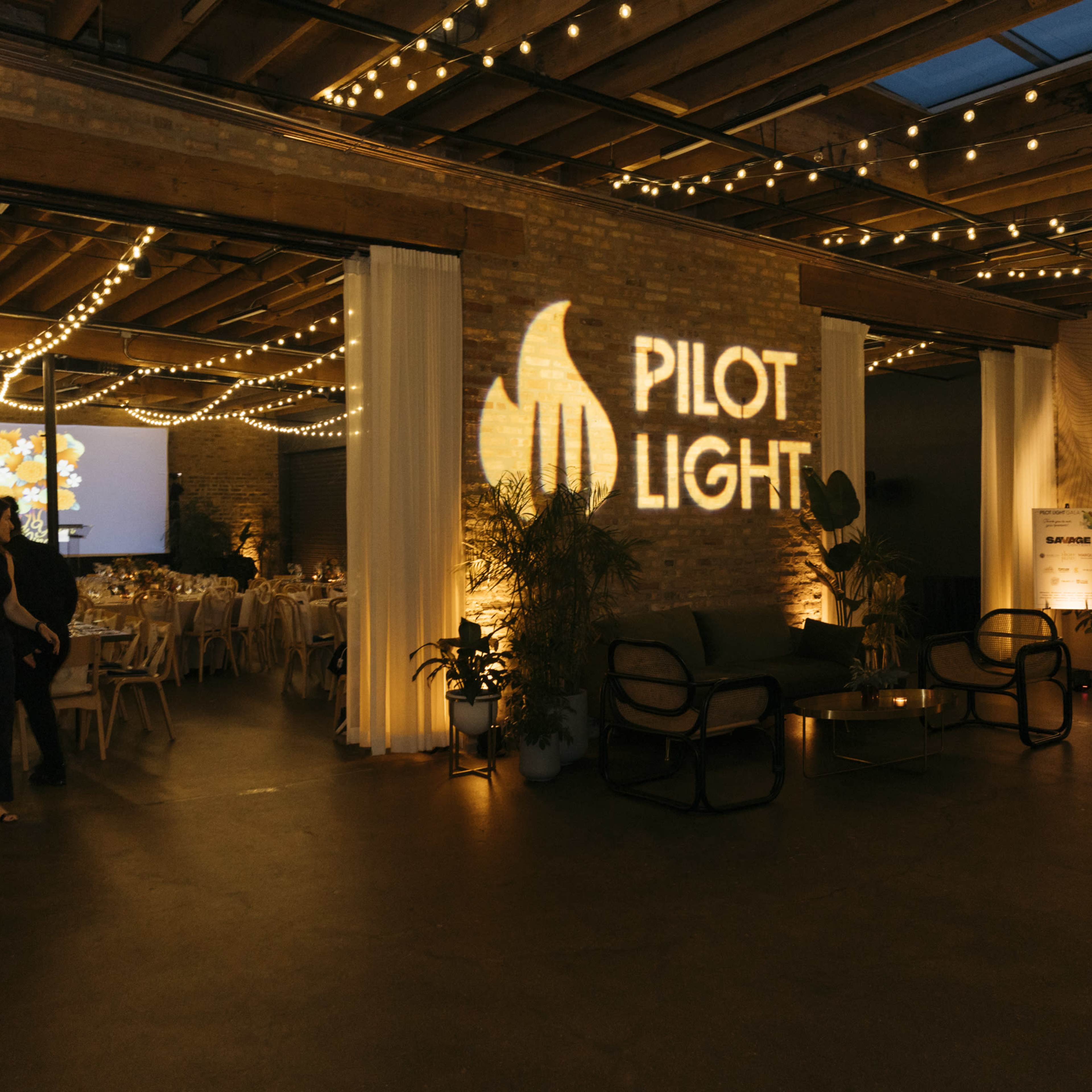 An event space with string lights, a projected logo reading "PILOT LIGHT," and arranged tables and chairs for a gathering.