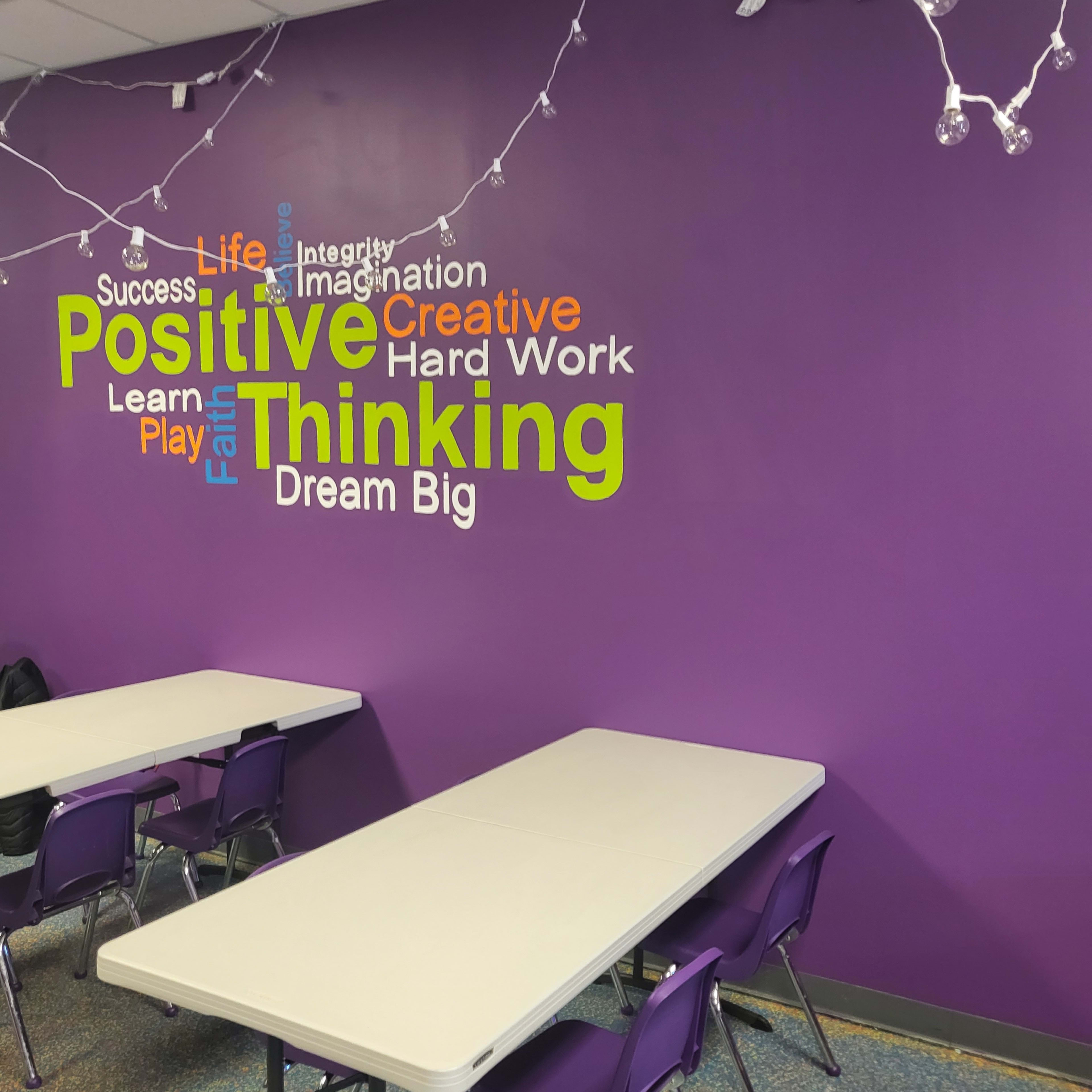 A room with purple walls featuring a word cloud of motivational phrases, alongside several tables and chairs set up for use.