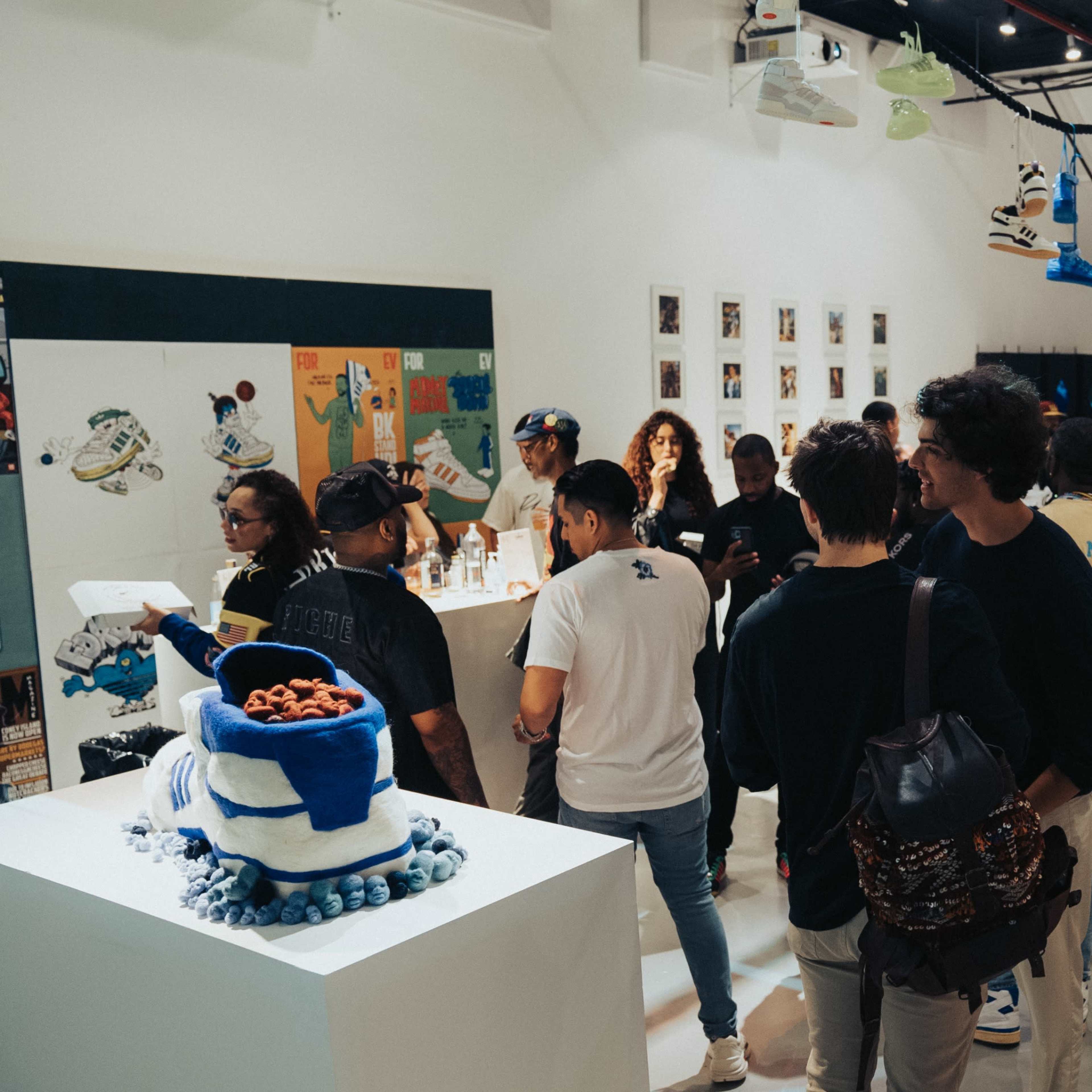 A crowded gallery exhibition features various artworks and displays related to sneakers, with attendees interacting and exploring the space.