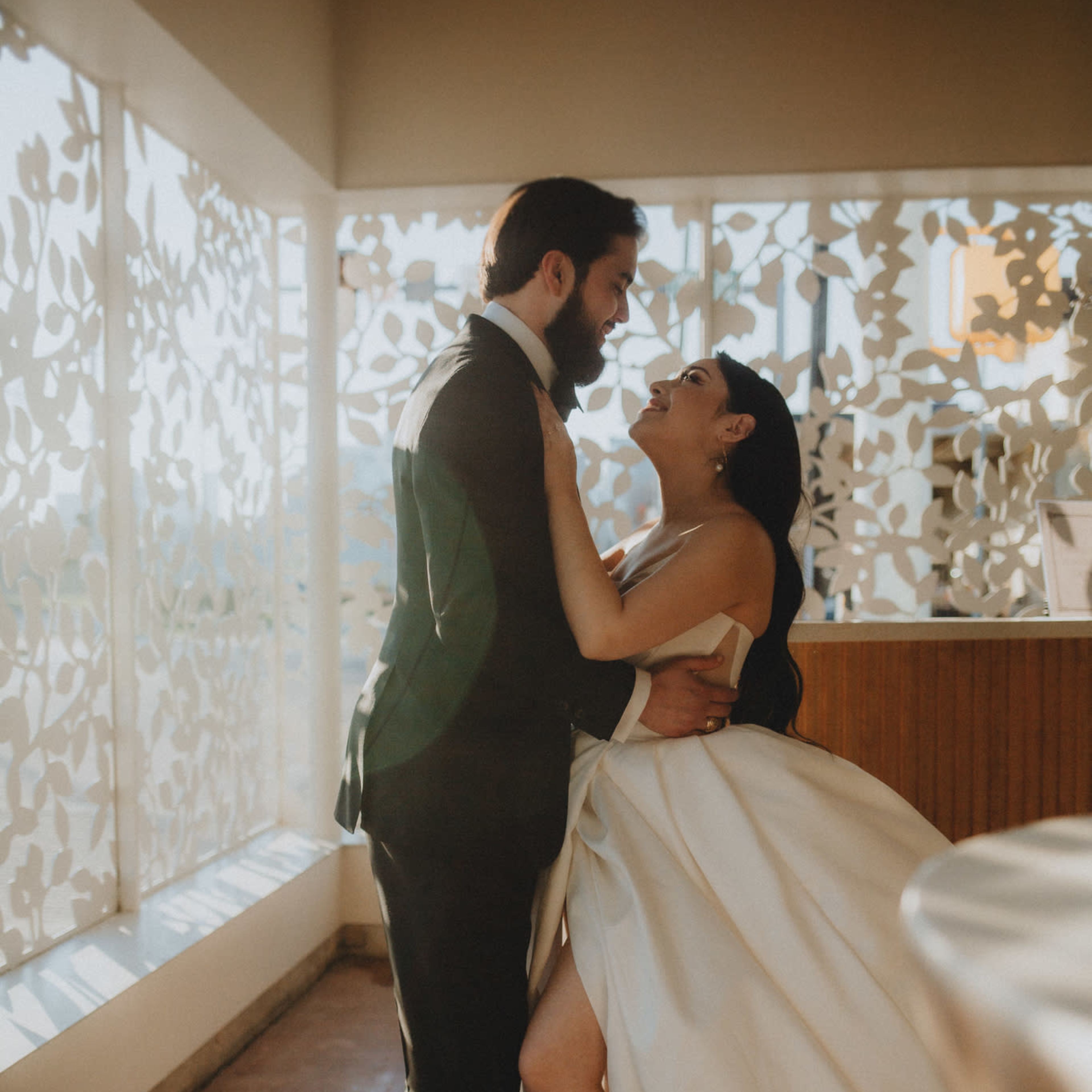 A bride and groom embrace warmly in a brightly lit room with decorative foliage-patterned windows.