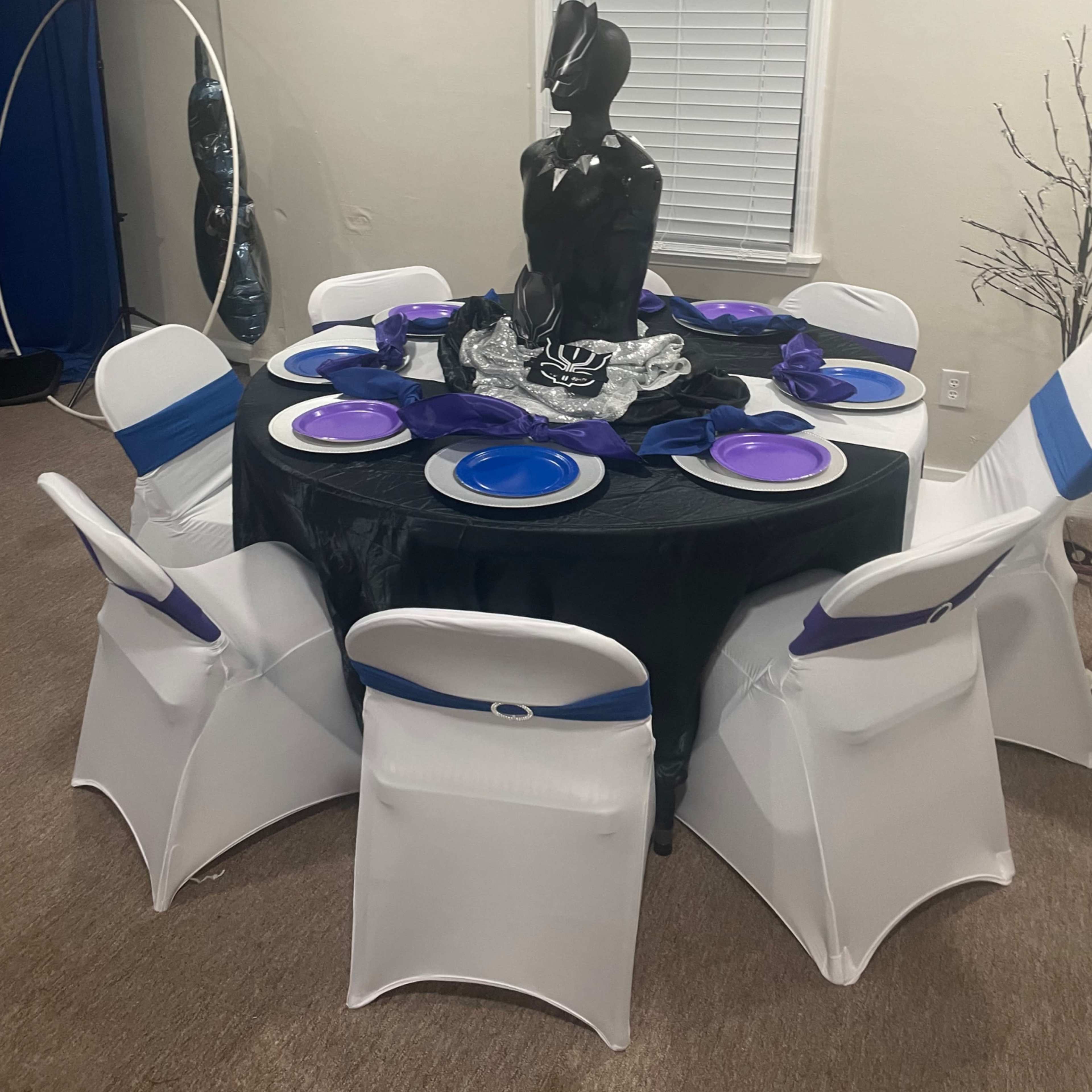 A round table with black and purple decorations is set with plates and a central black statue surrounded by six chairs.