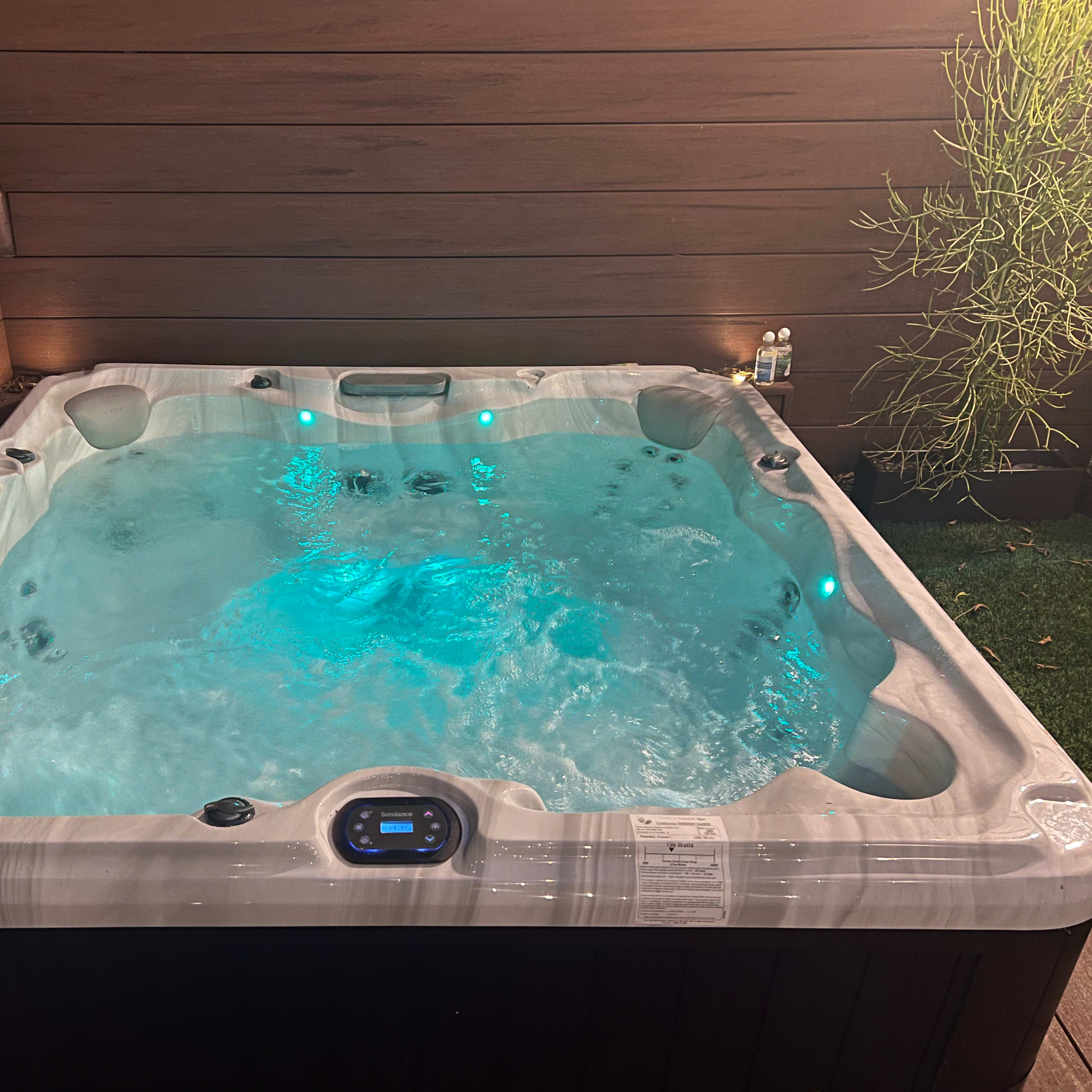 A rectangular hot tub with blue underwater lighting is positioned against a wooden wall in a landscaped area.