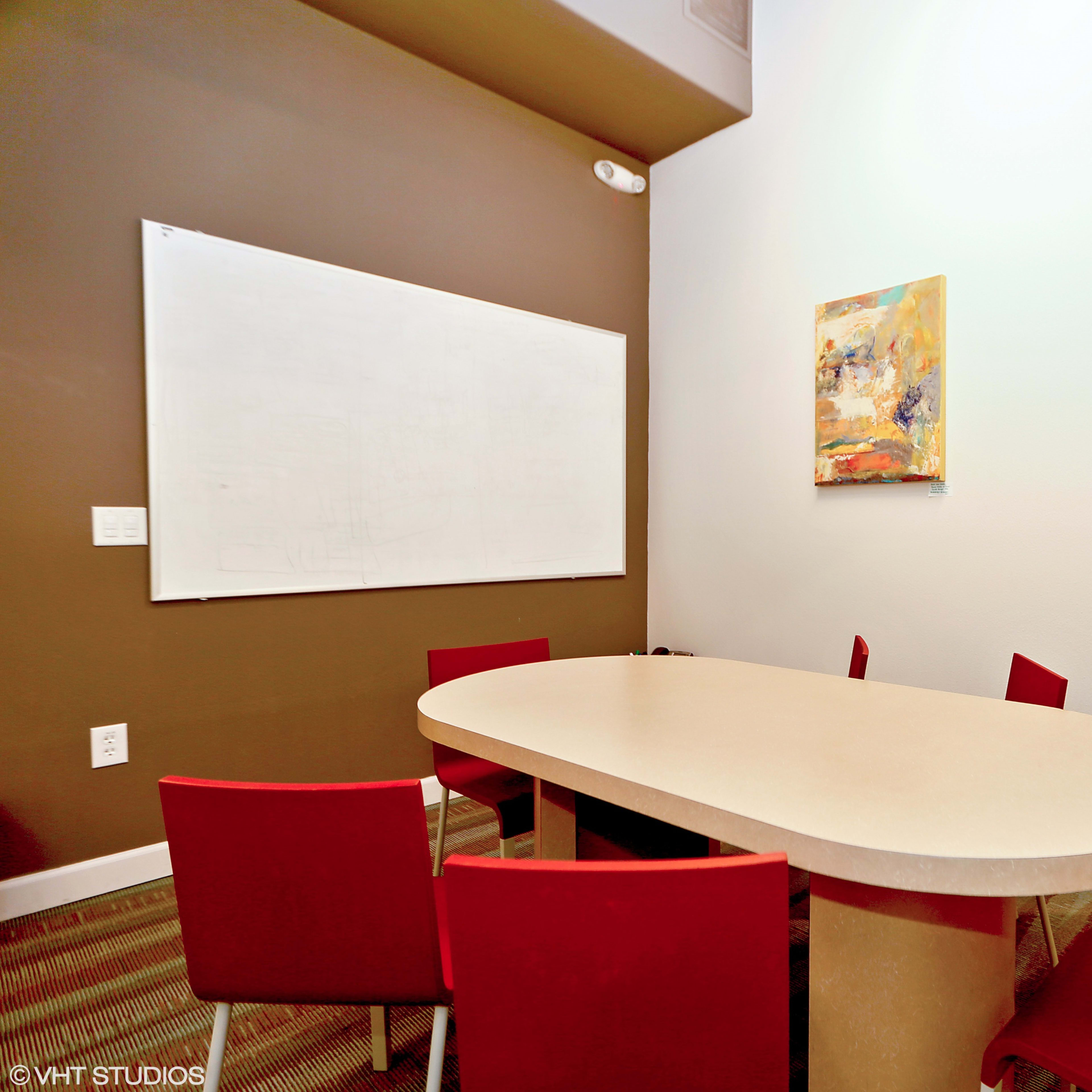 A small conference room features a large oval table surrounded by red chairs and a whiteboard on one wall.
