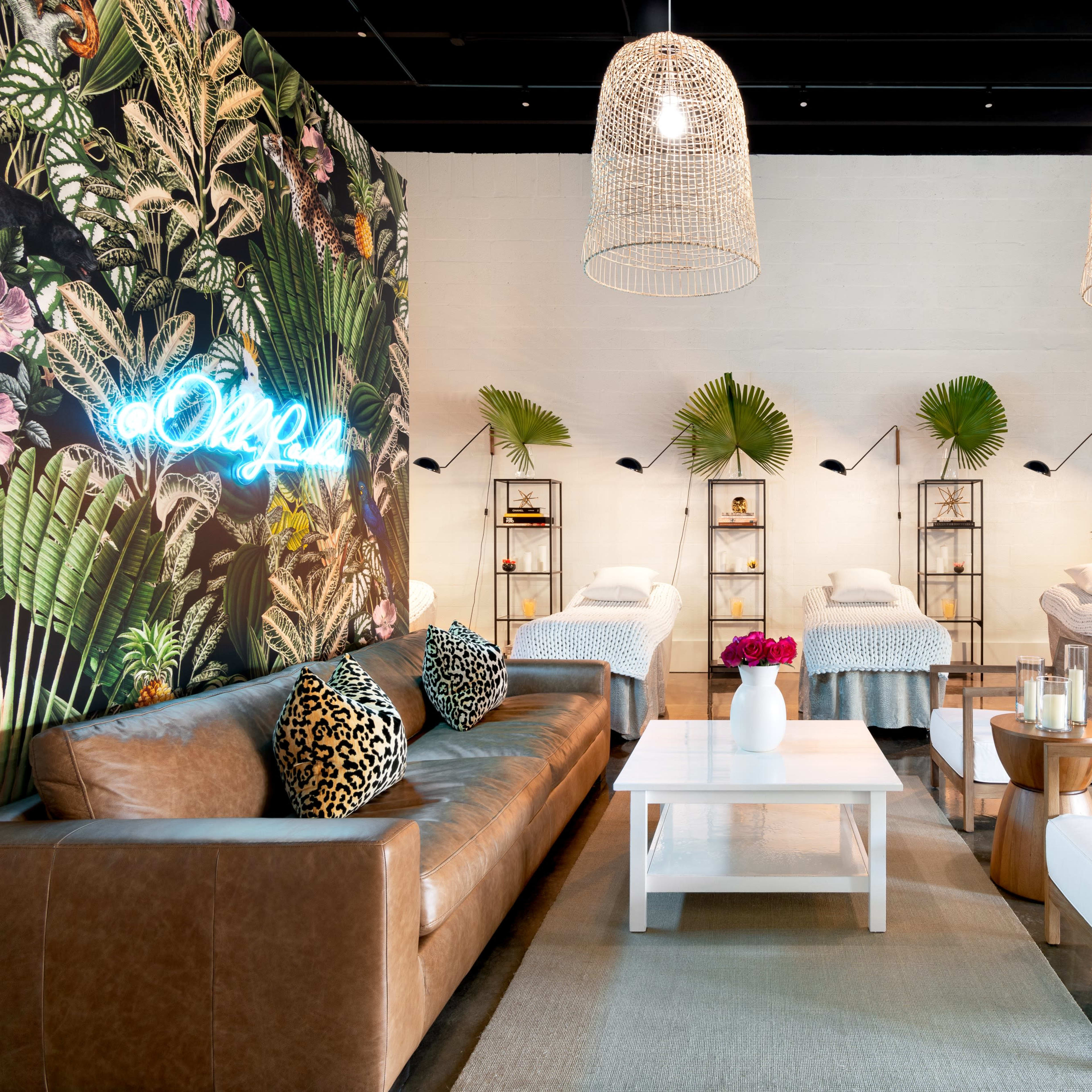 A modern spa interior featuring a leafy accent wall, several treatment beds arranged in a line, and stylish seating areas with pendant light fixtures overhead.