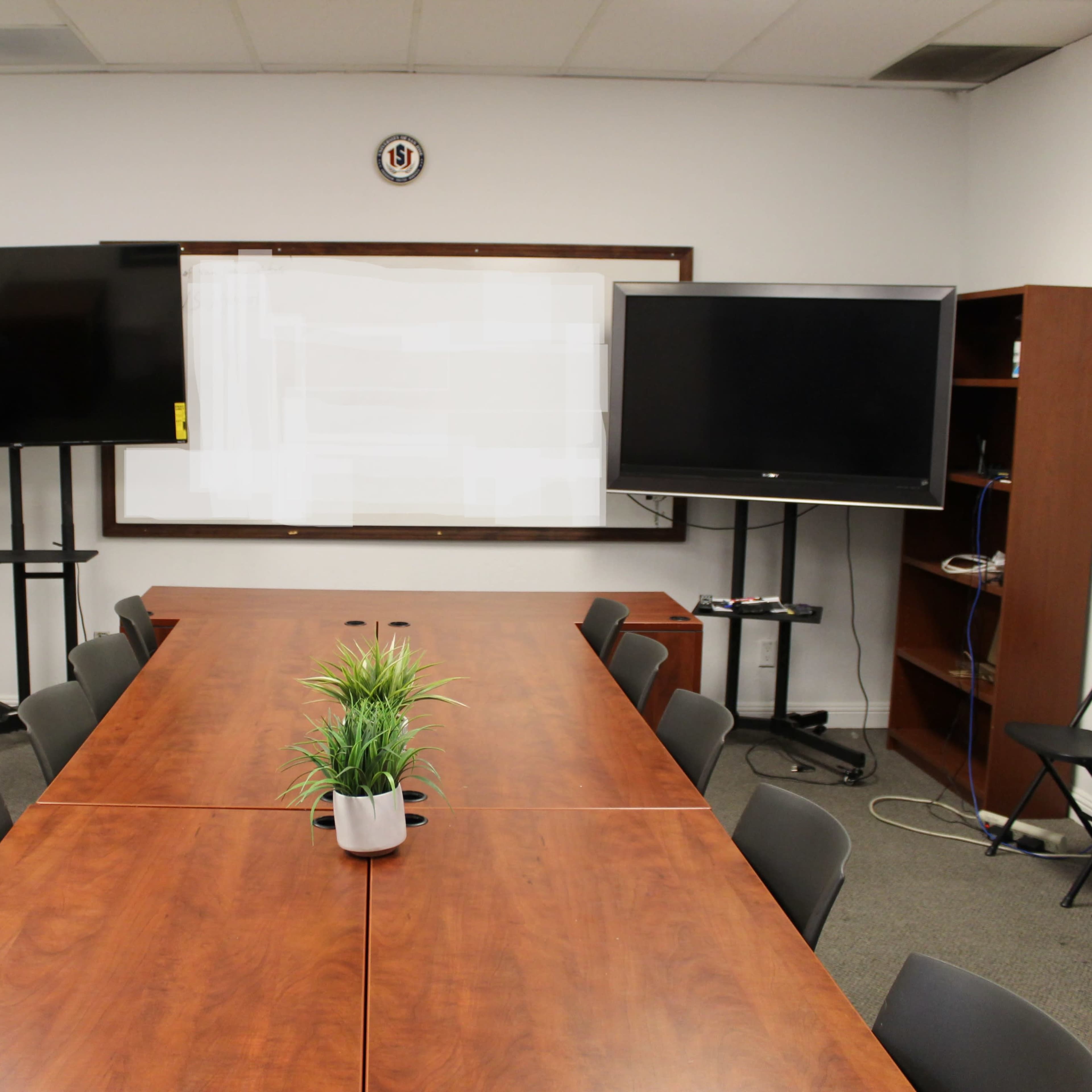 A conference room features a long wooden table with a small plant, flanked by two television screens and a whiteboard at the front.