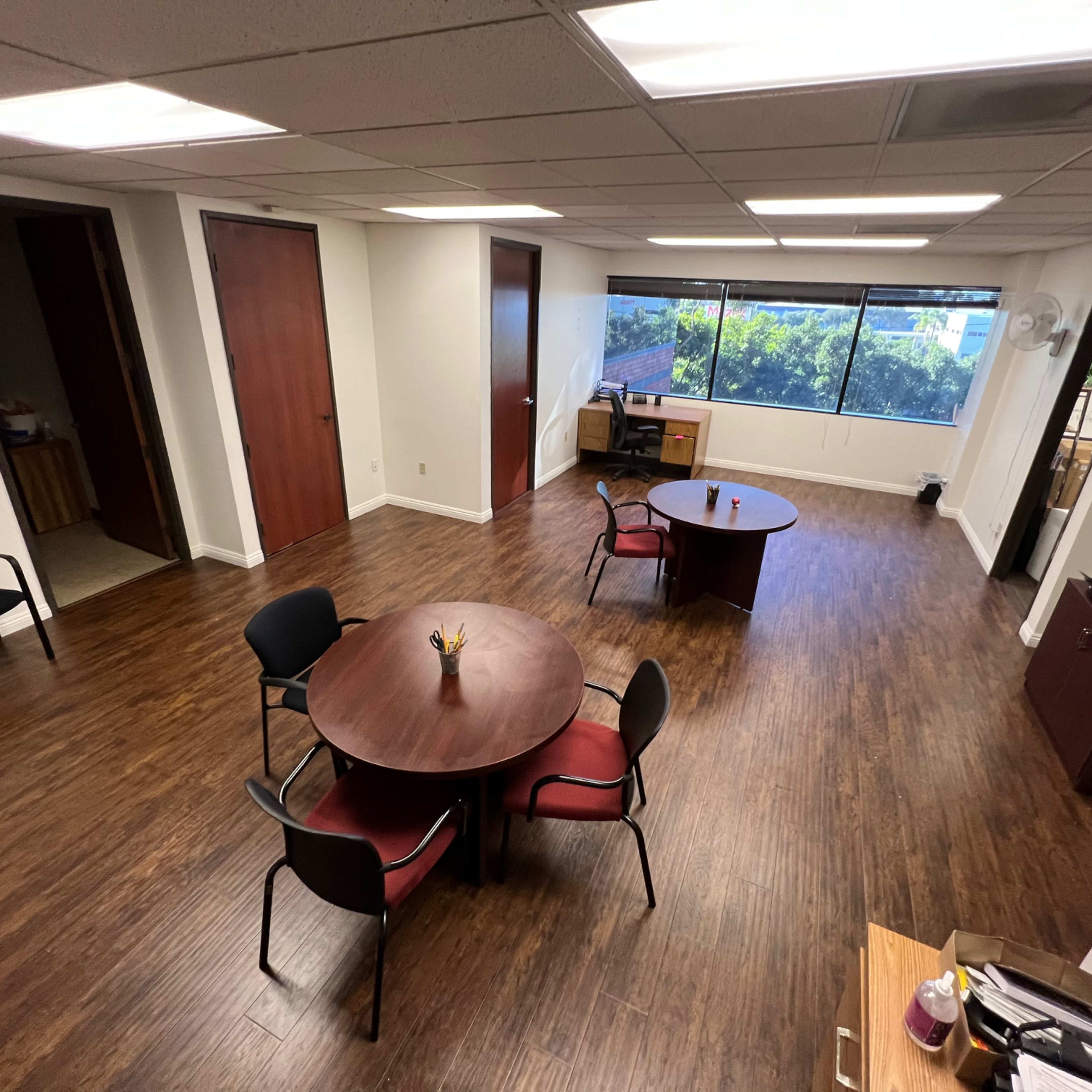 An office space featuring two round tables with chairs, a desk near a window, and wooden flooring.