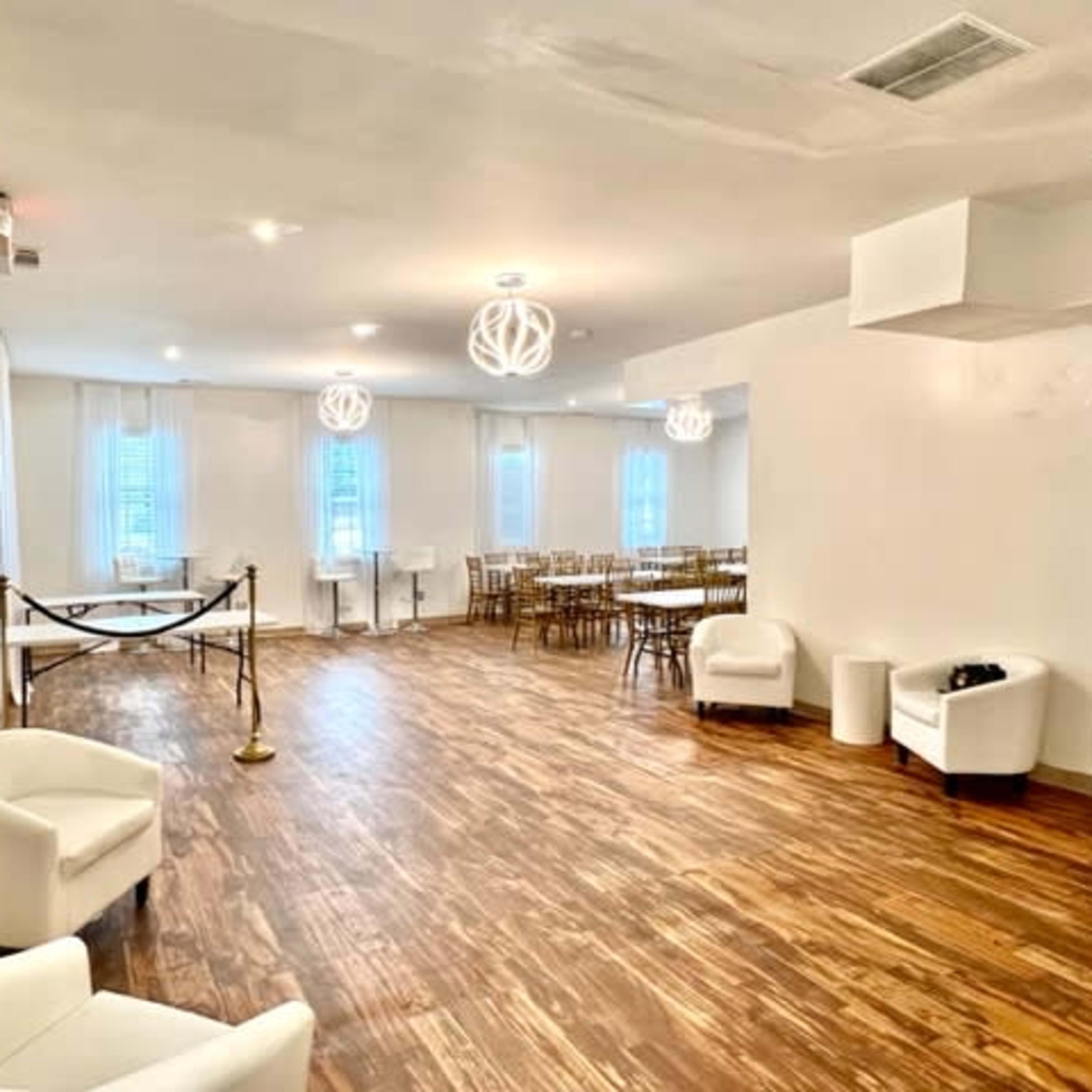 The interior of a spacious, well-lit venue features wooden flooring, white walls, and a setup of tables and chairs in a dining area.