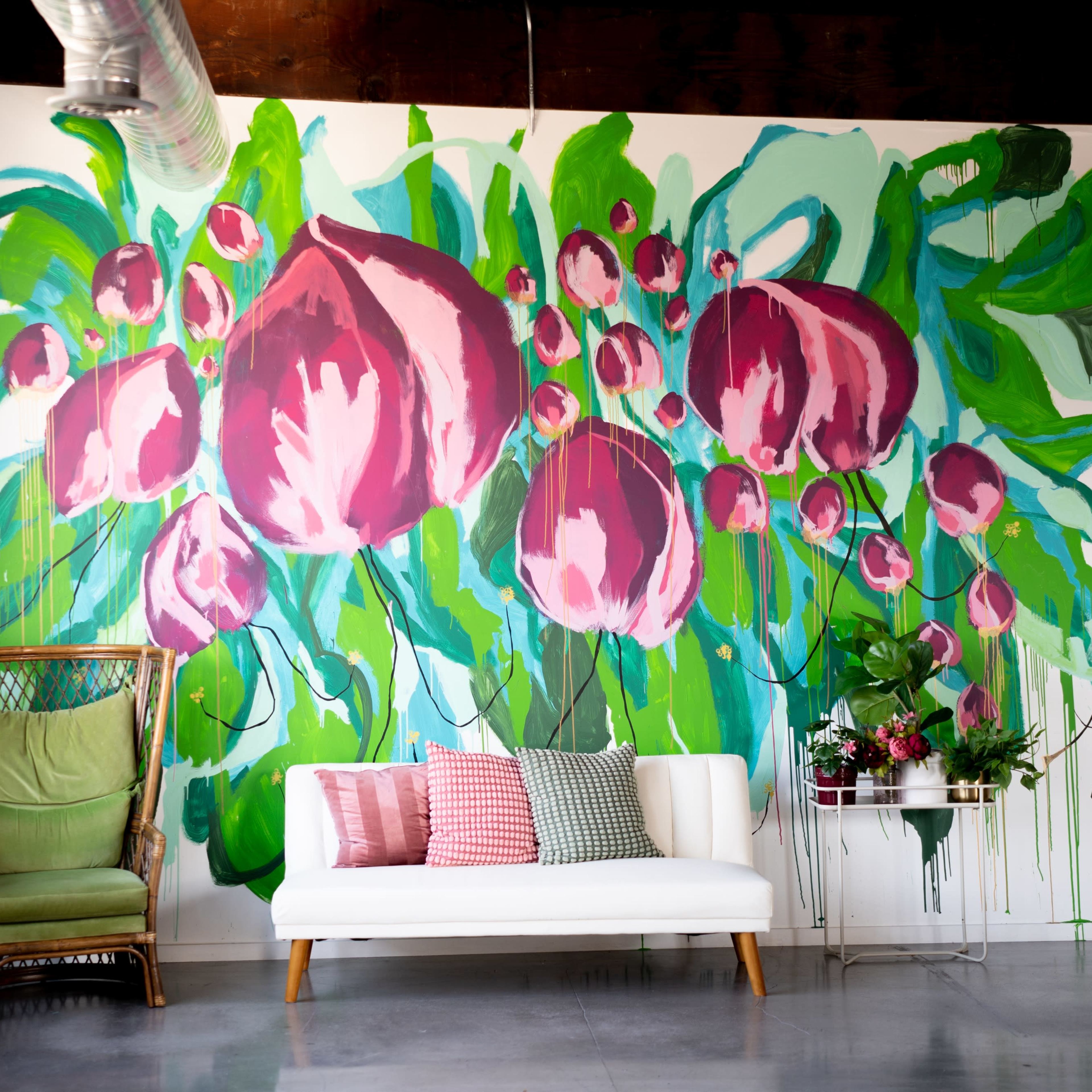 A bright interior with a large floral mural on the wall, a white sofa, a colorful pillow arrangement, and a green chair.