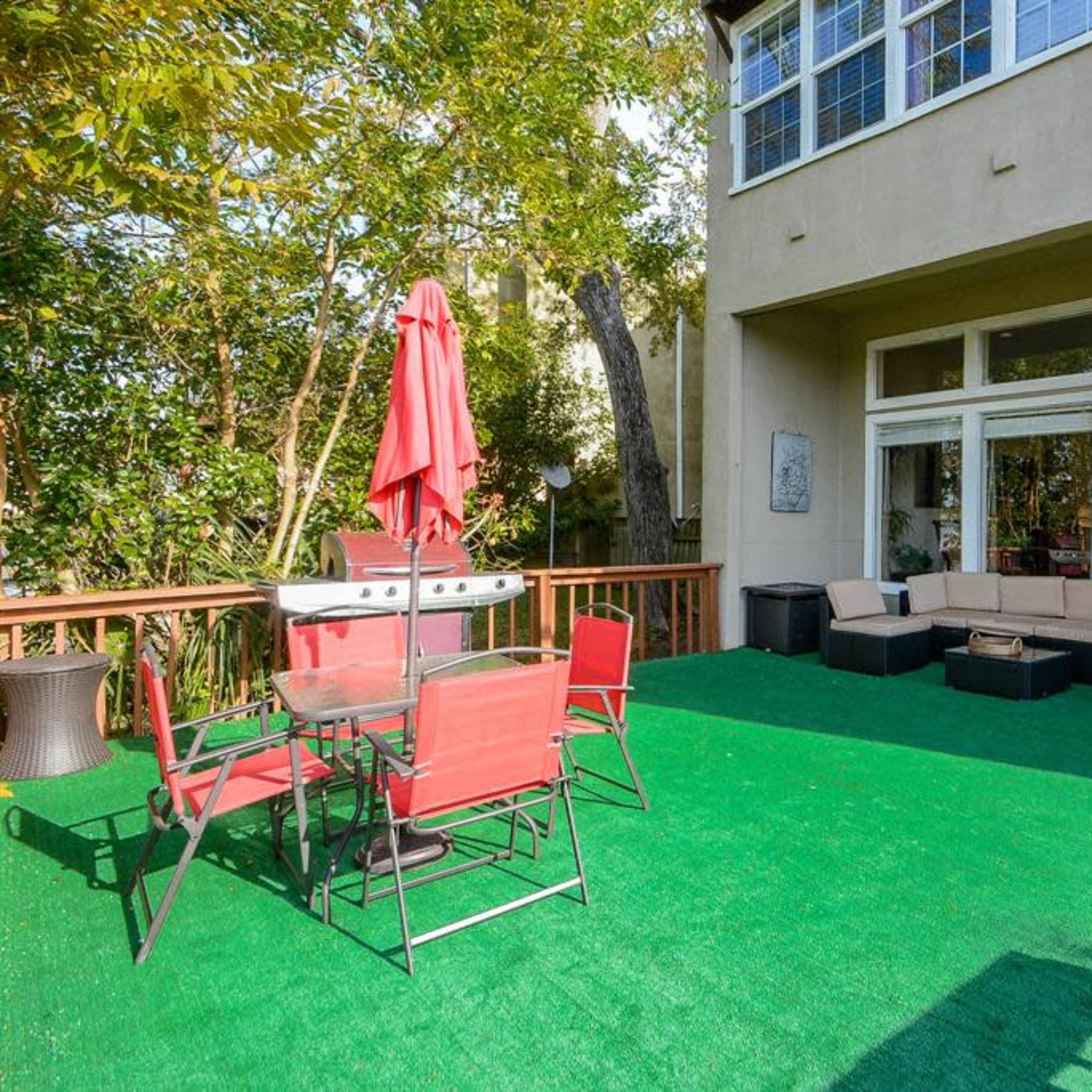 A backyard deck area with a seating arrangement, a dining table with red chairs, a grill, and artificial grass.