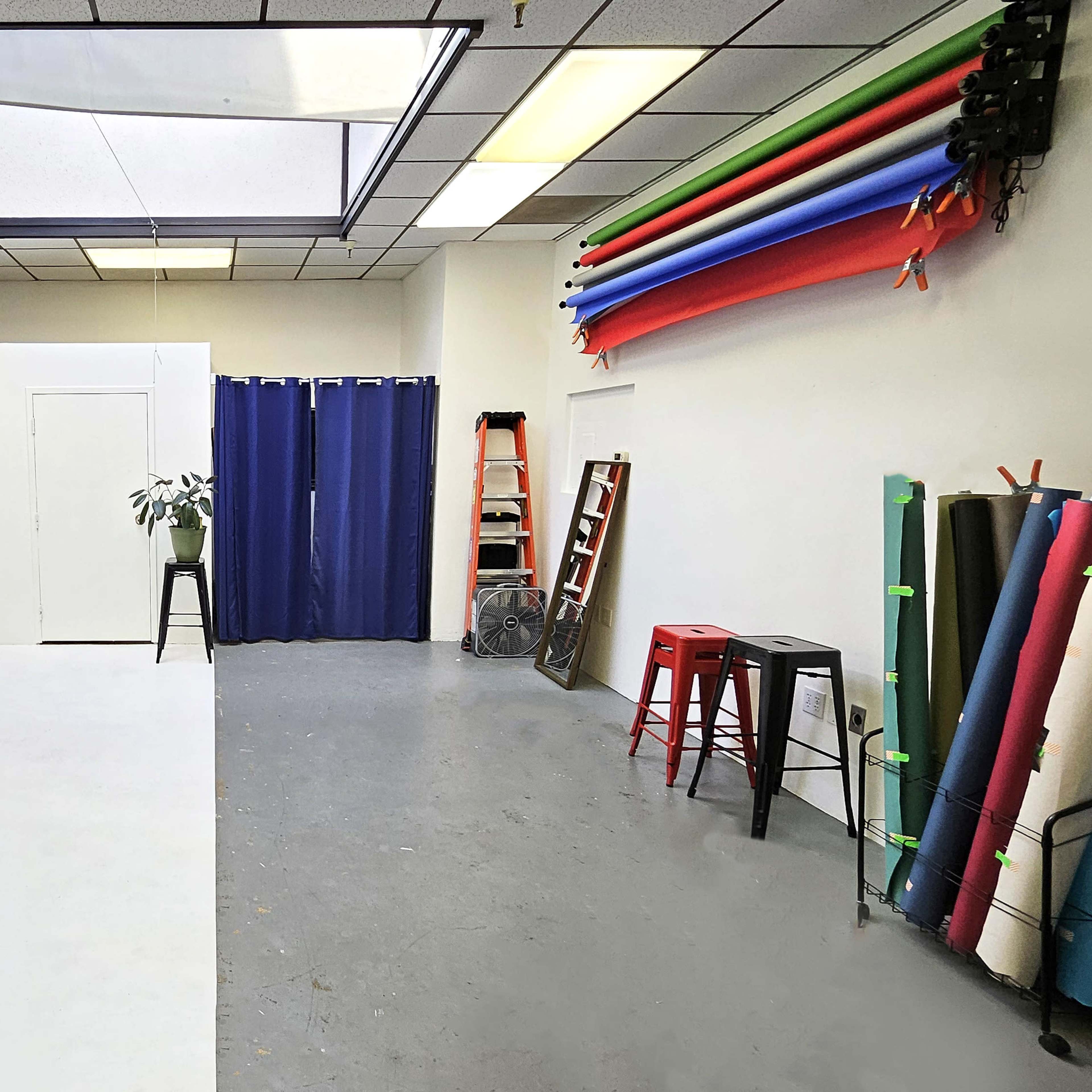 A studio space with a white backdrop, a large skylight, colorful backdrop rolls on the walls, and a few stools and a ladder positioned in the room.