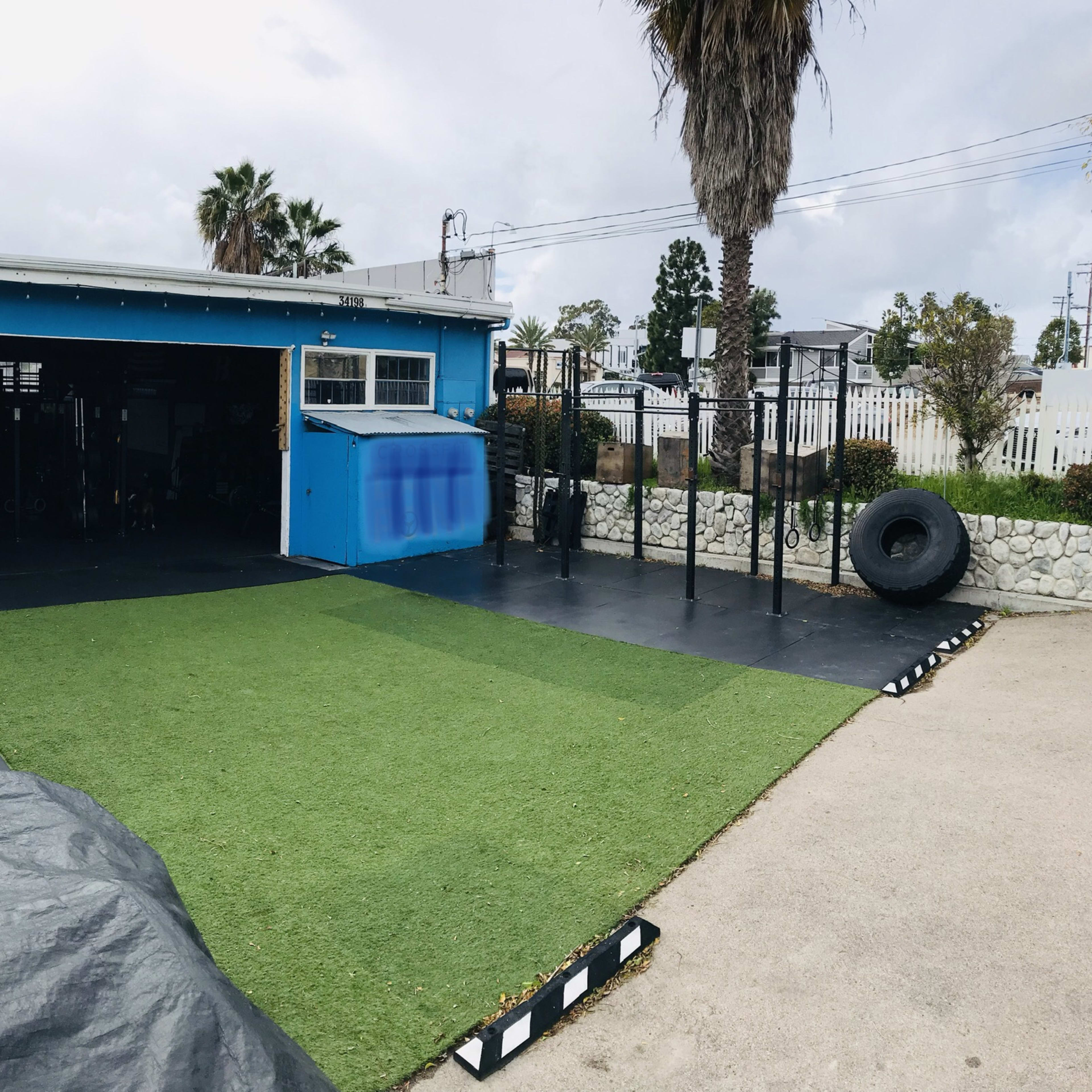 An outdoor gym area featuring turf flooring, exercise equipment, and a blue building in the background.