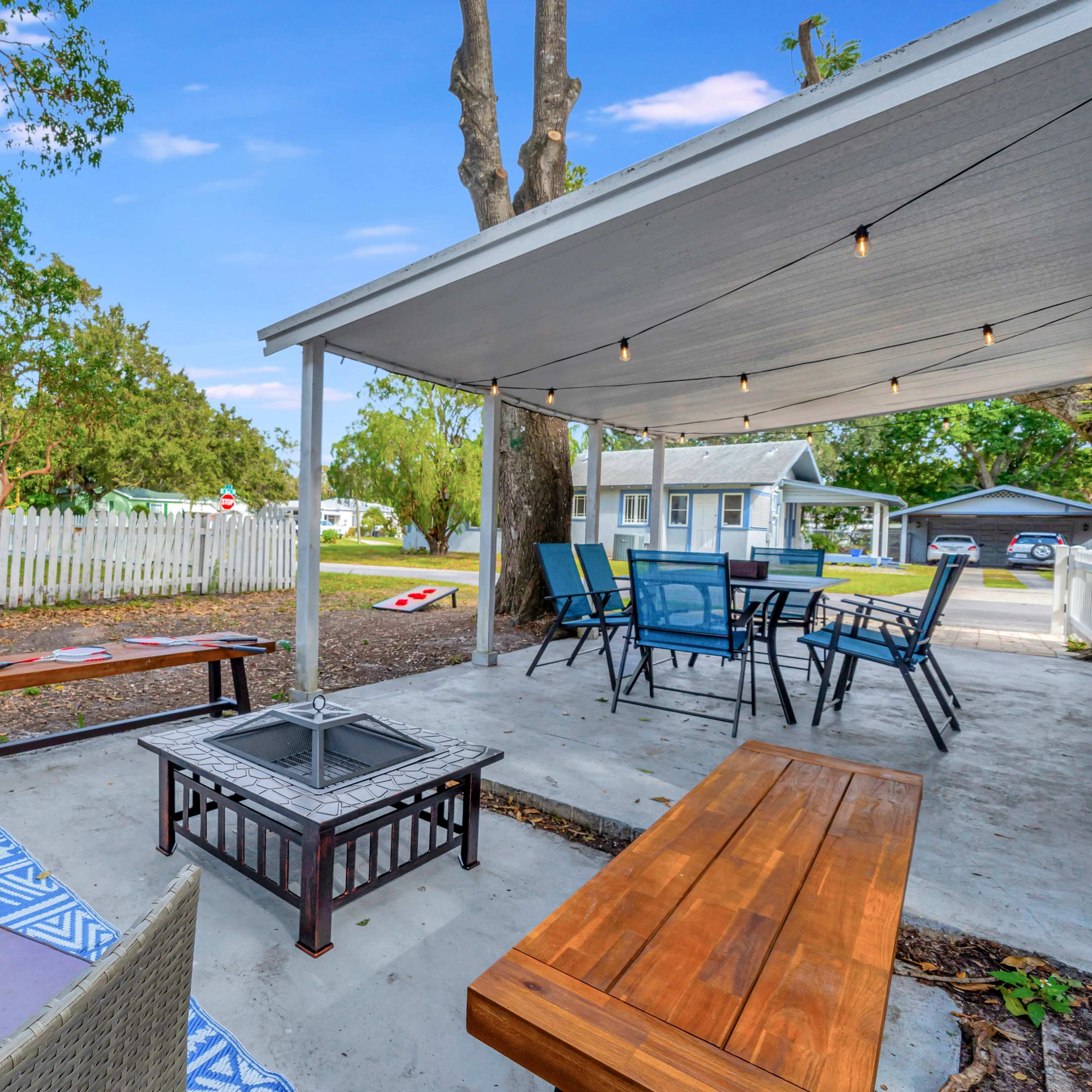 A patio area with outdoor seating, a fire pit, and string lights beneath a roofed structure, surrounded by trees and a white fence.