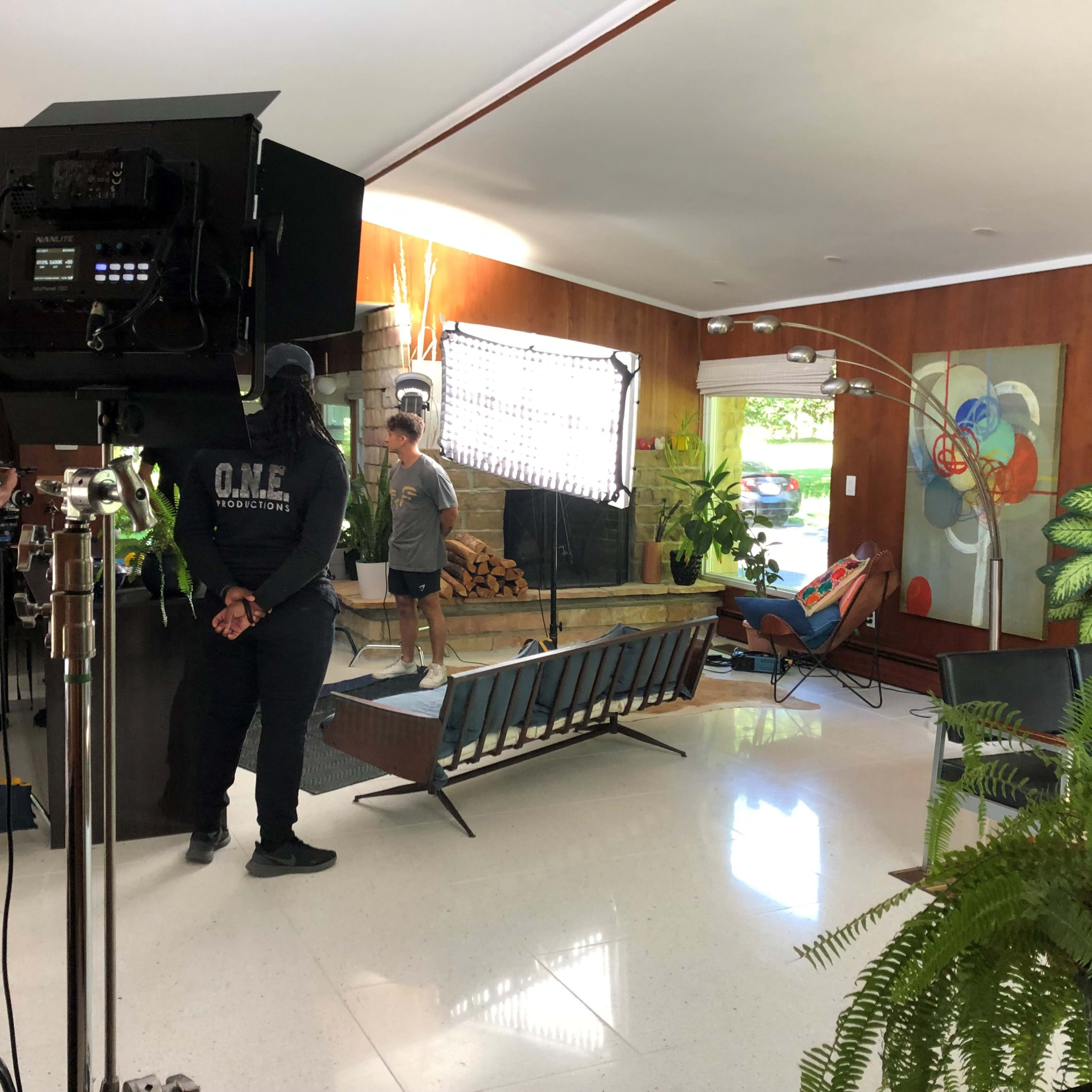 A film crew sets up lighting equipment in a modern living room with wooden paneling and plants.