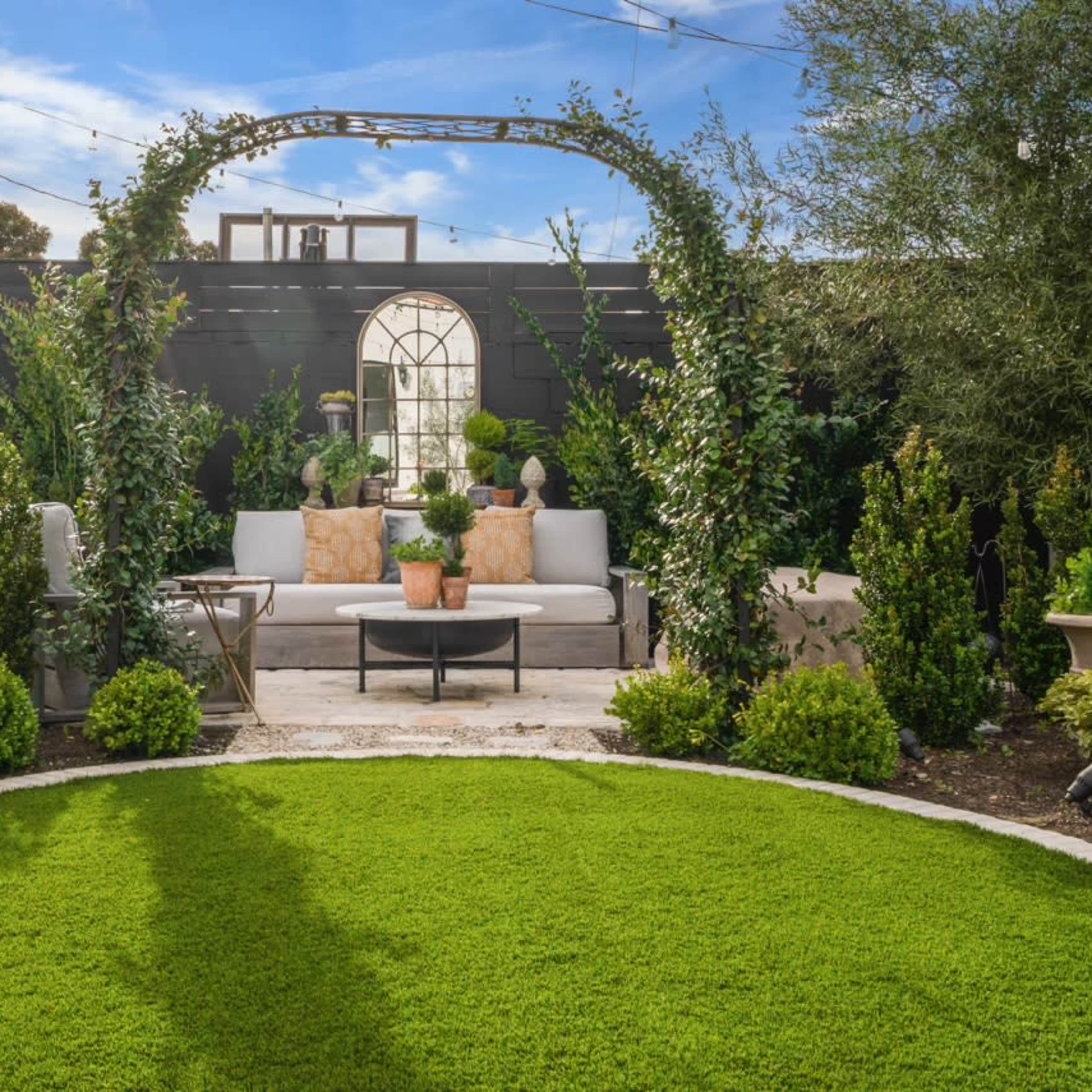 A landscaped garden featuring a curved stone seating area surrounded by greenery and an archway adorned with plants.