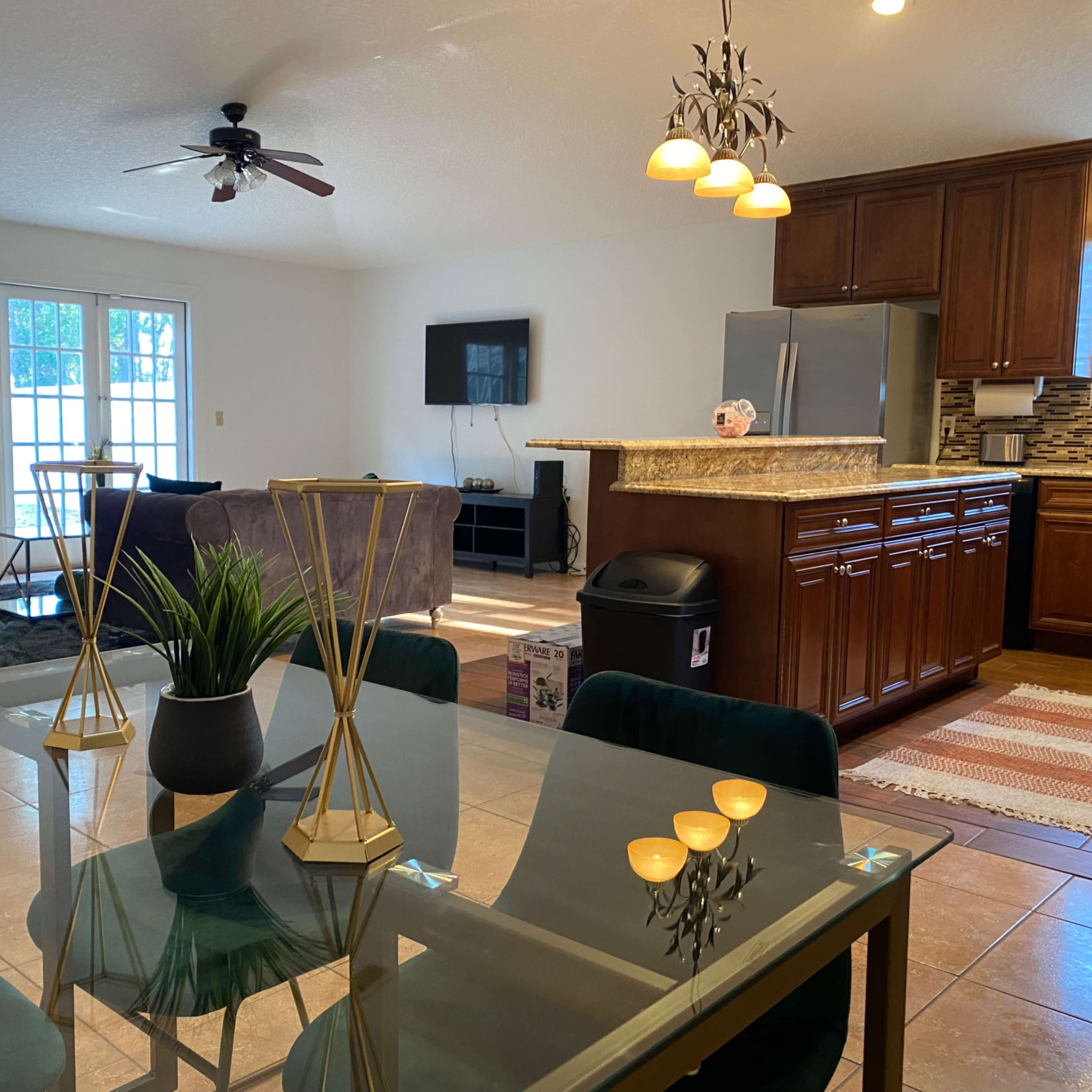 A spacious living and dining area features a glass dining table, a kitchen with wooden cabinets, and a seating area with plush couches.