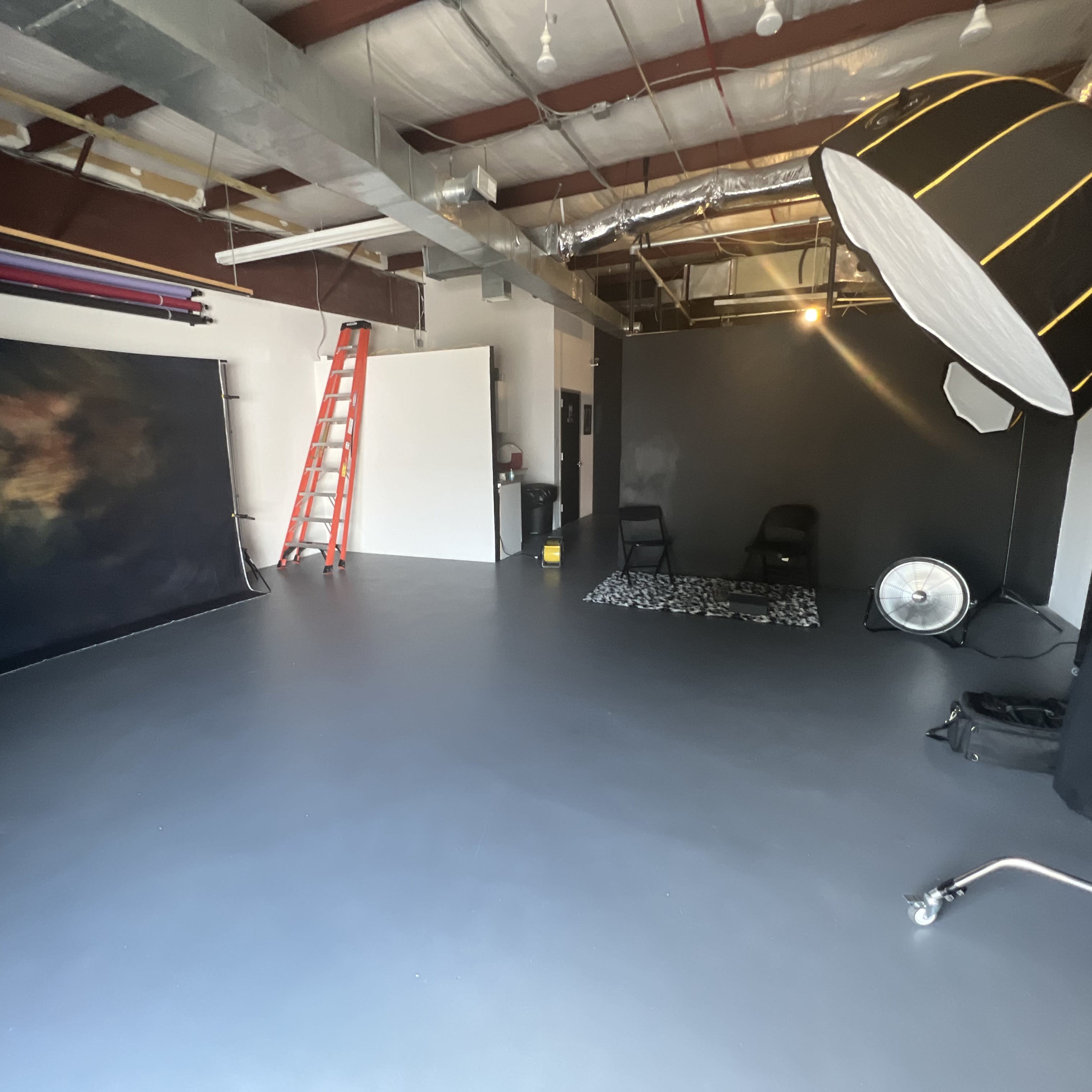 A spacious photography studio with a ladder, backdrop, lighting equipment, and a fan.