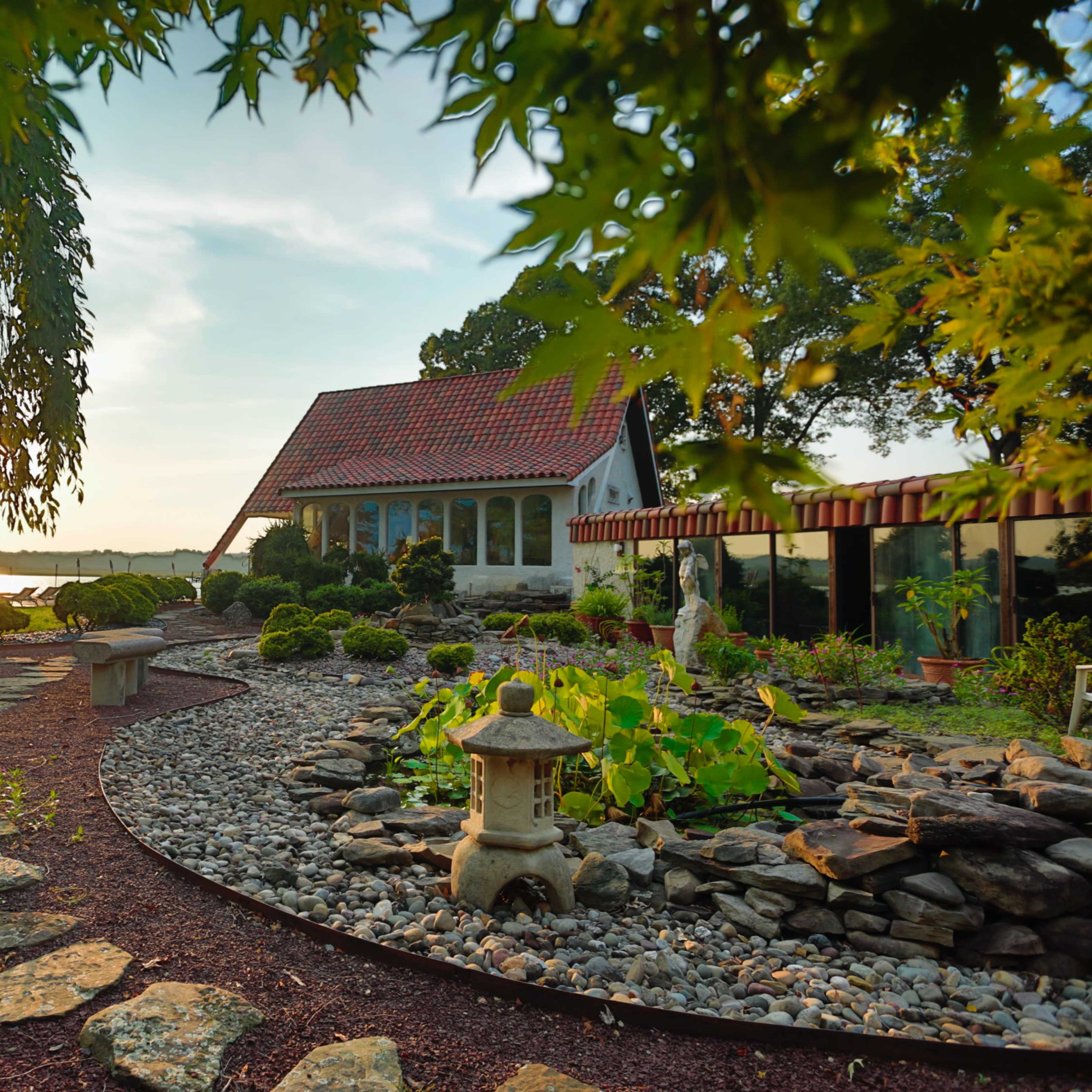 A garden path winds through a landscaped area with a stone lantern and a view of a house near a body of water.