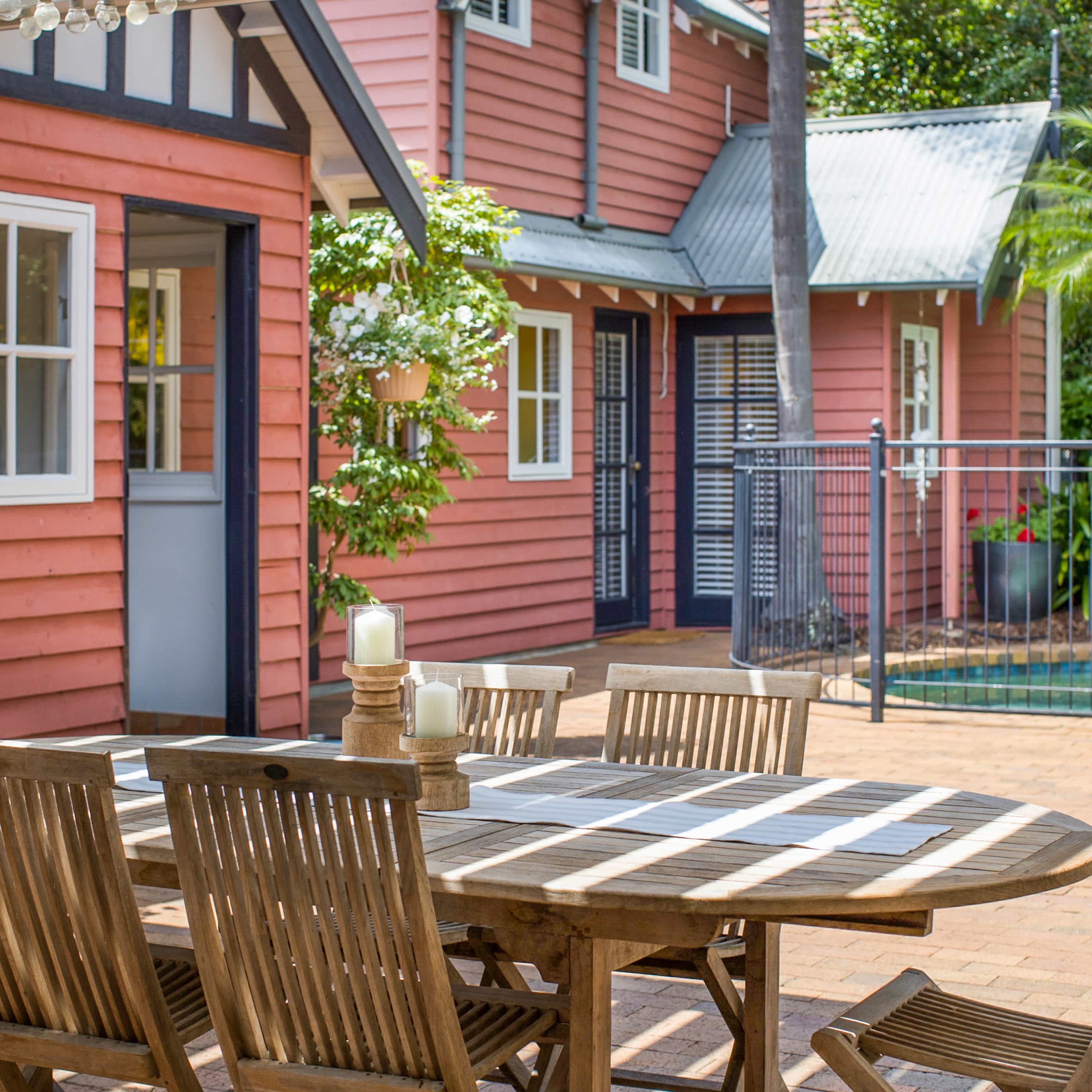 A wooden dining table with chairs is set up on a patio next to a pink house and a swimming pool surrounded by greenery.