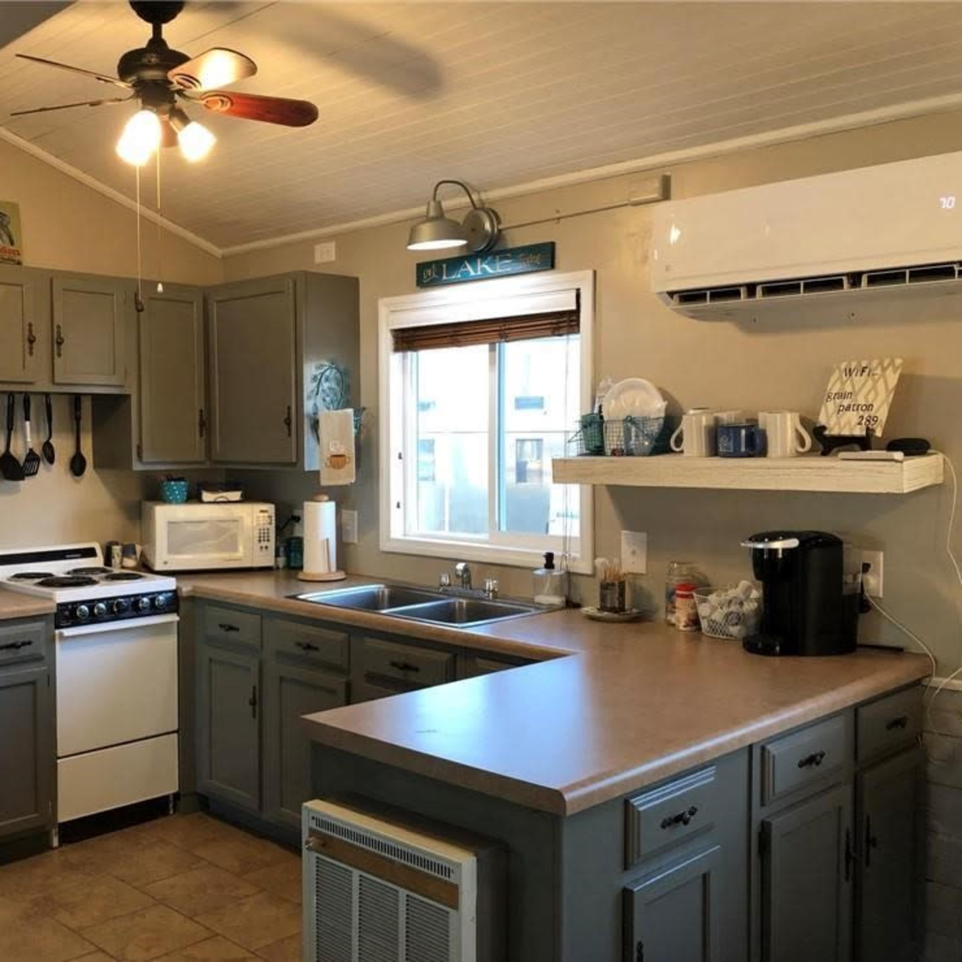 A compact kitchen with gray cabinets, a white refrigerator, a stove, a microwave, and a sink, illuminated by a ceiling fan and natural light from a window.