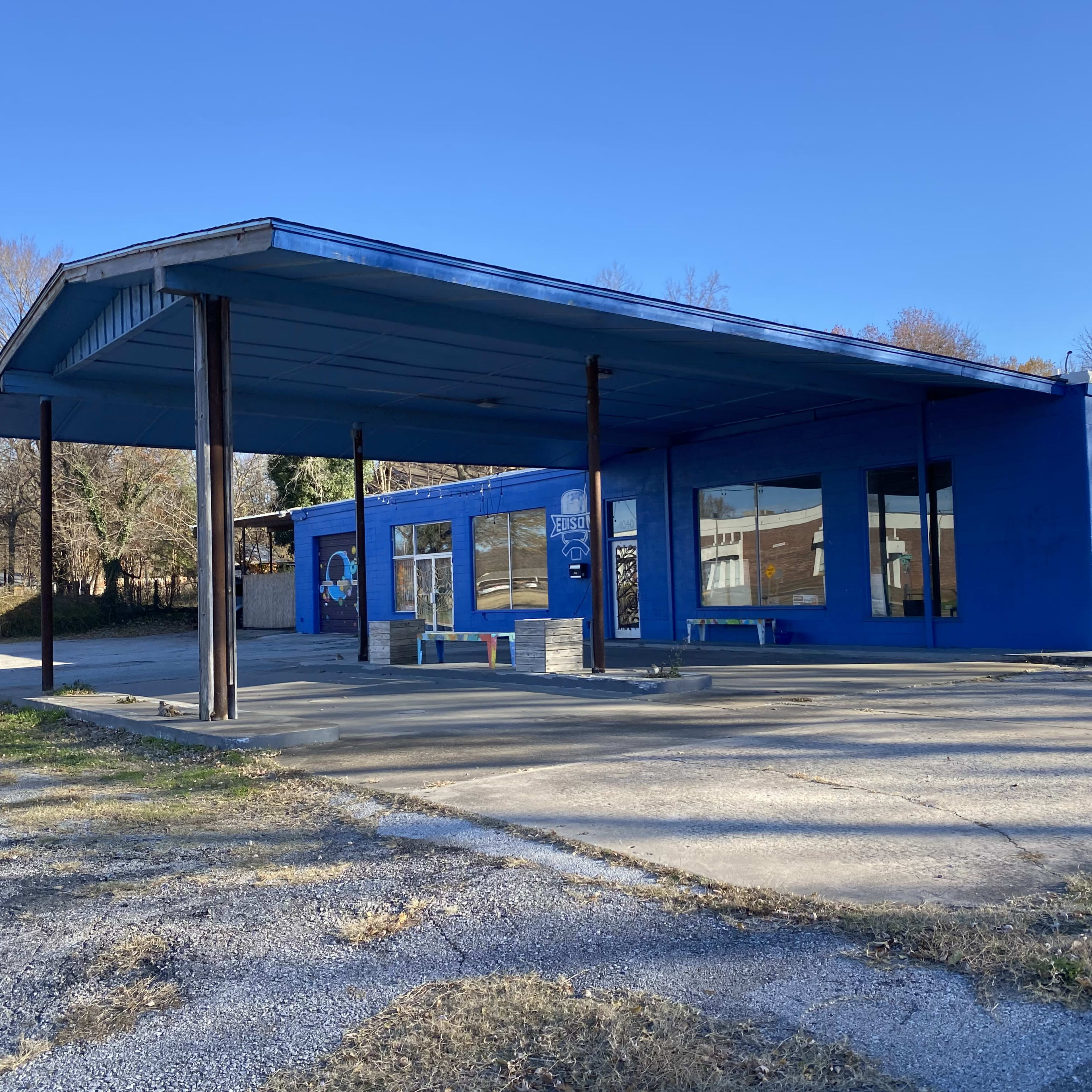 A blue-painted building with a canopy and large windows stands on a gravel lot, surrounded by trees.