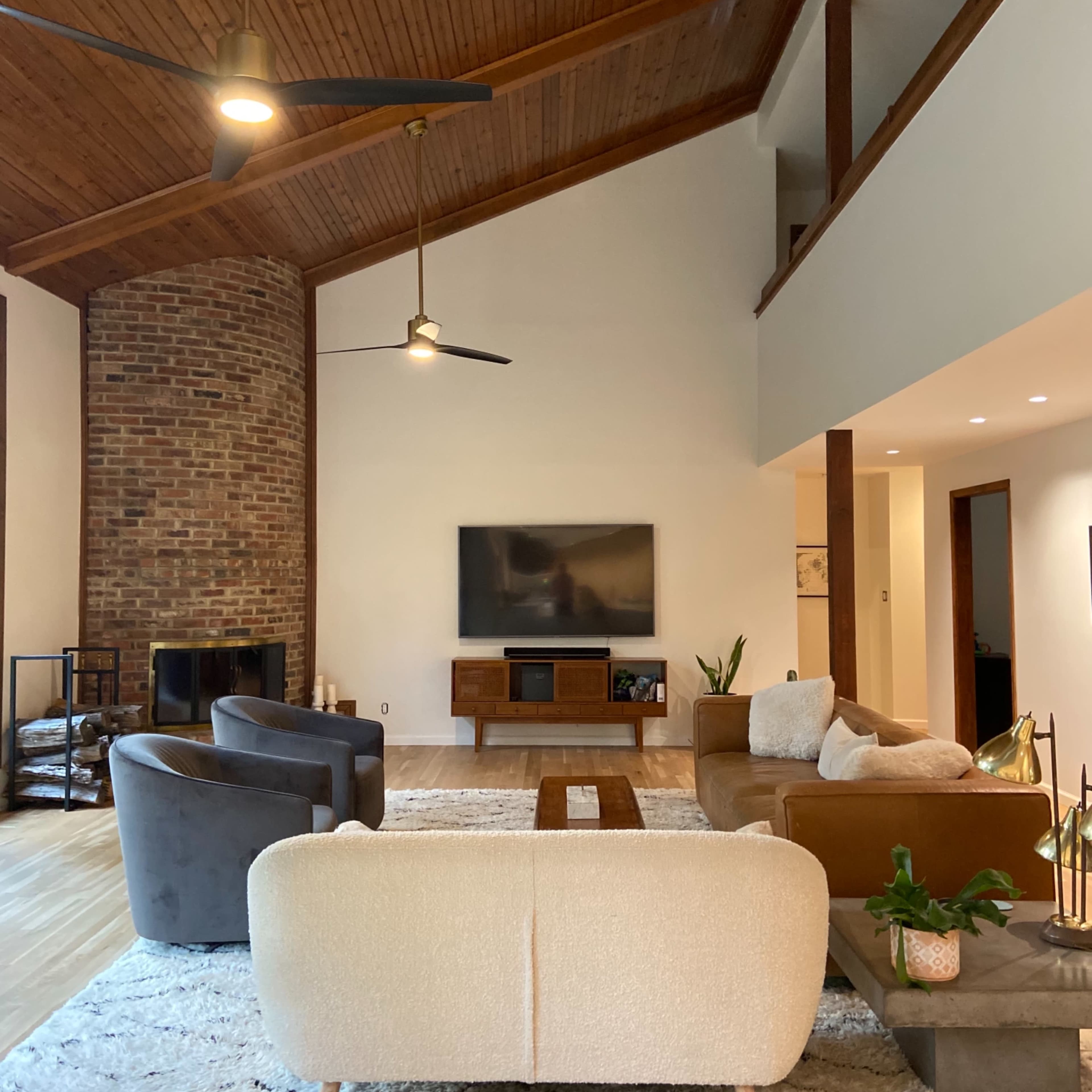 A spacious living room with a high wooden ceiling, a brick fireplace, a large television on the wall, and a mix of modern furniture arranged on a light area rug.