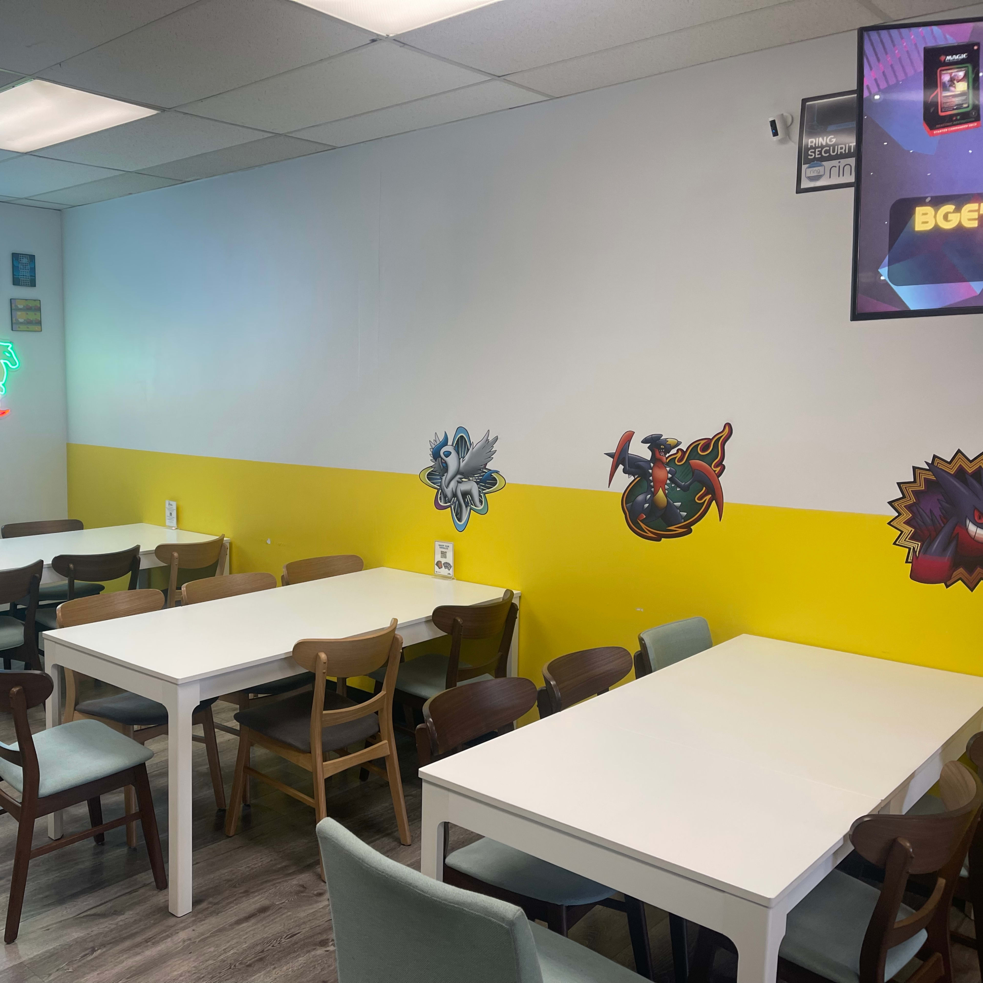 A brightly colored game room with white tables, wooden chairs, and wall decorations featuring characters and a neon sign that reads "GAME ON."