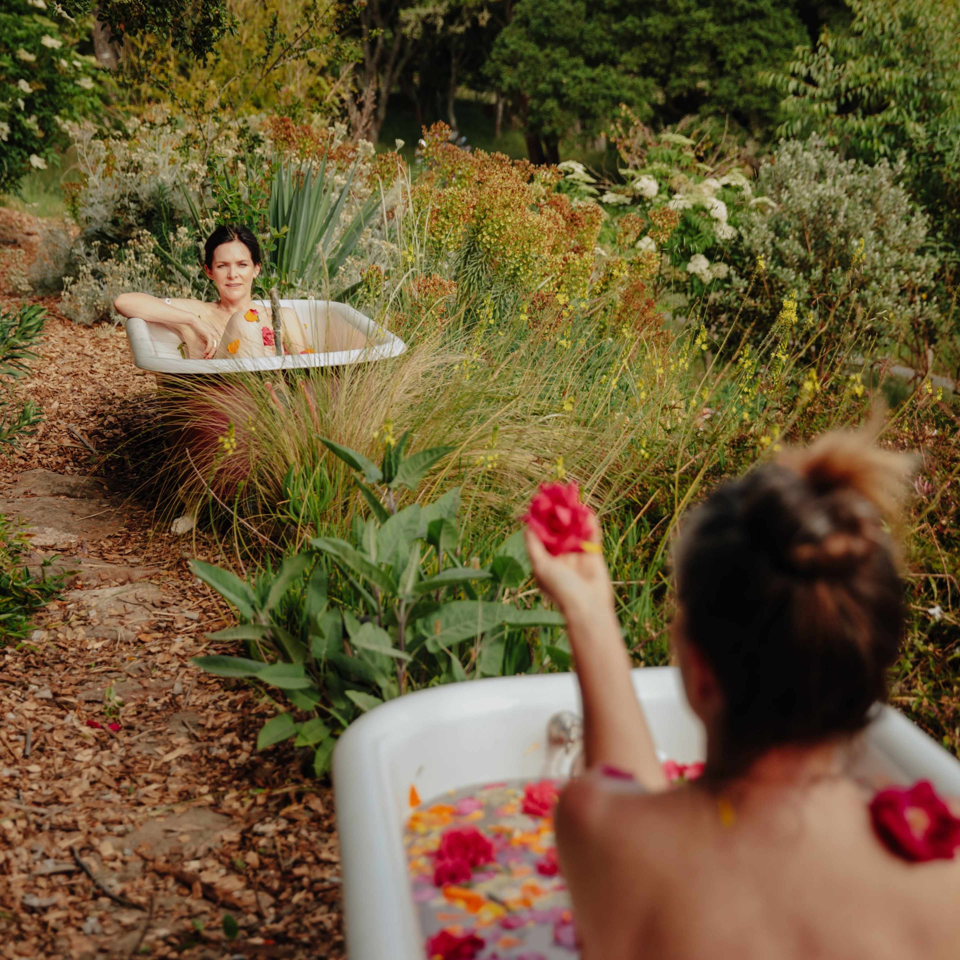 Two people are sitting in bathtubs placed outdoors among lush greenery and flowers, each holding a flower.