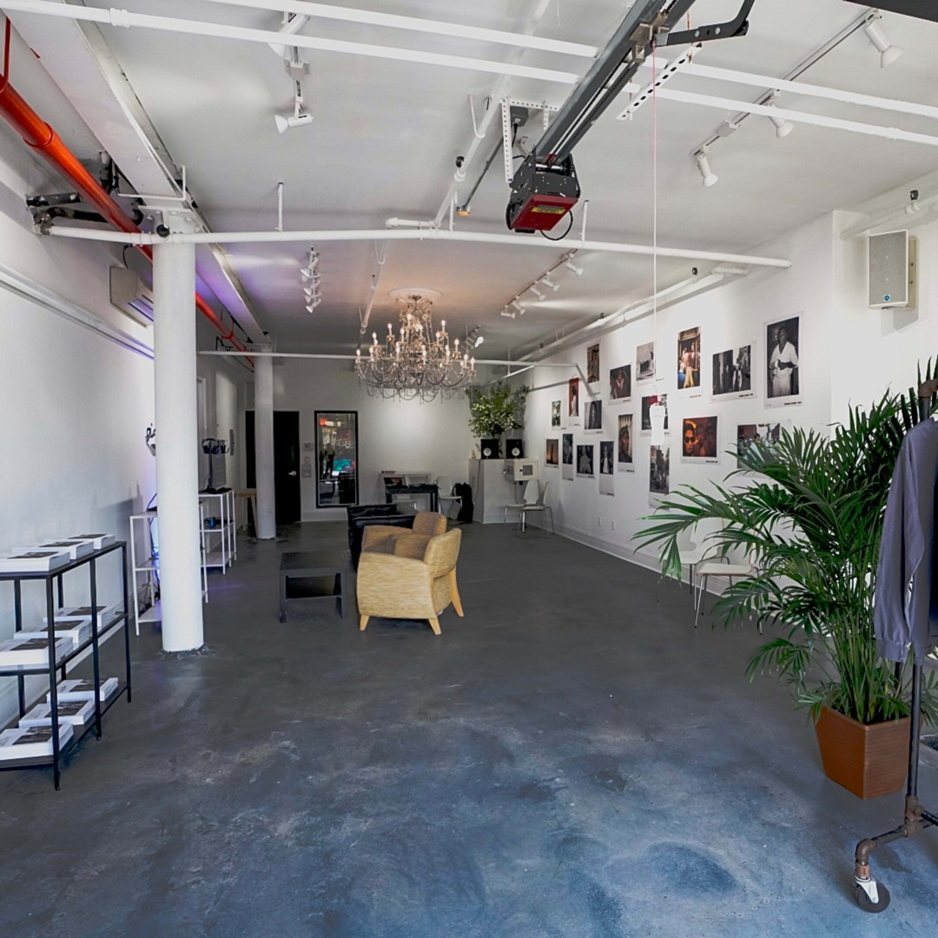 A spacious, well-lit gallery or studio with a chandelier, seating area, and clothing displayed on racks along one wall.