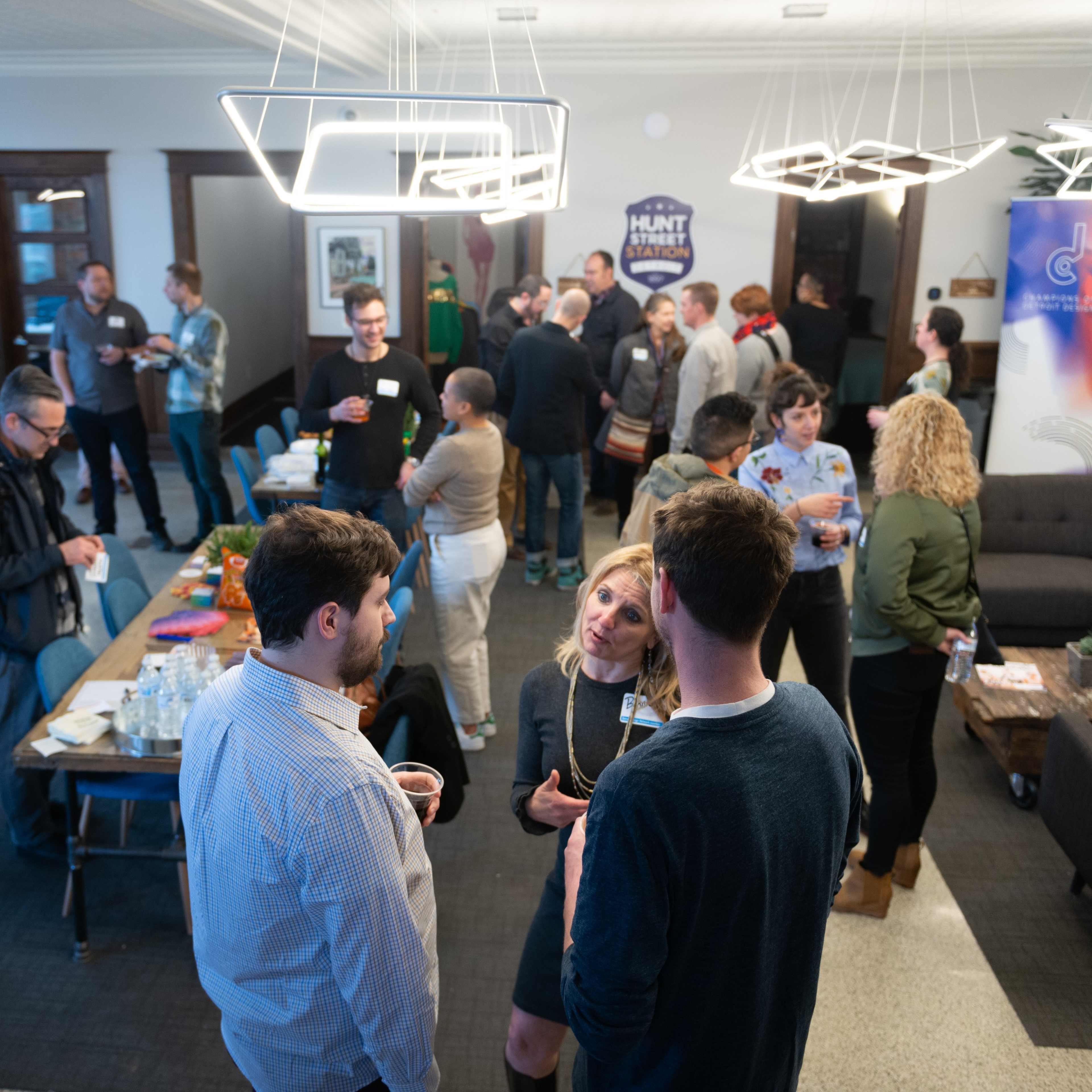 A group of people socializes at a networking event in a modern indoor space with a lounge area and refreshments laid out on a table.