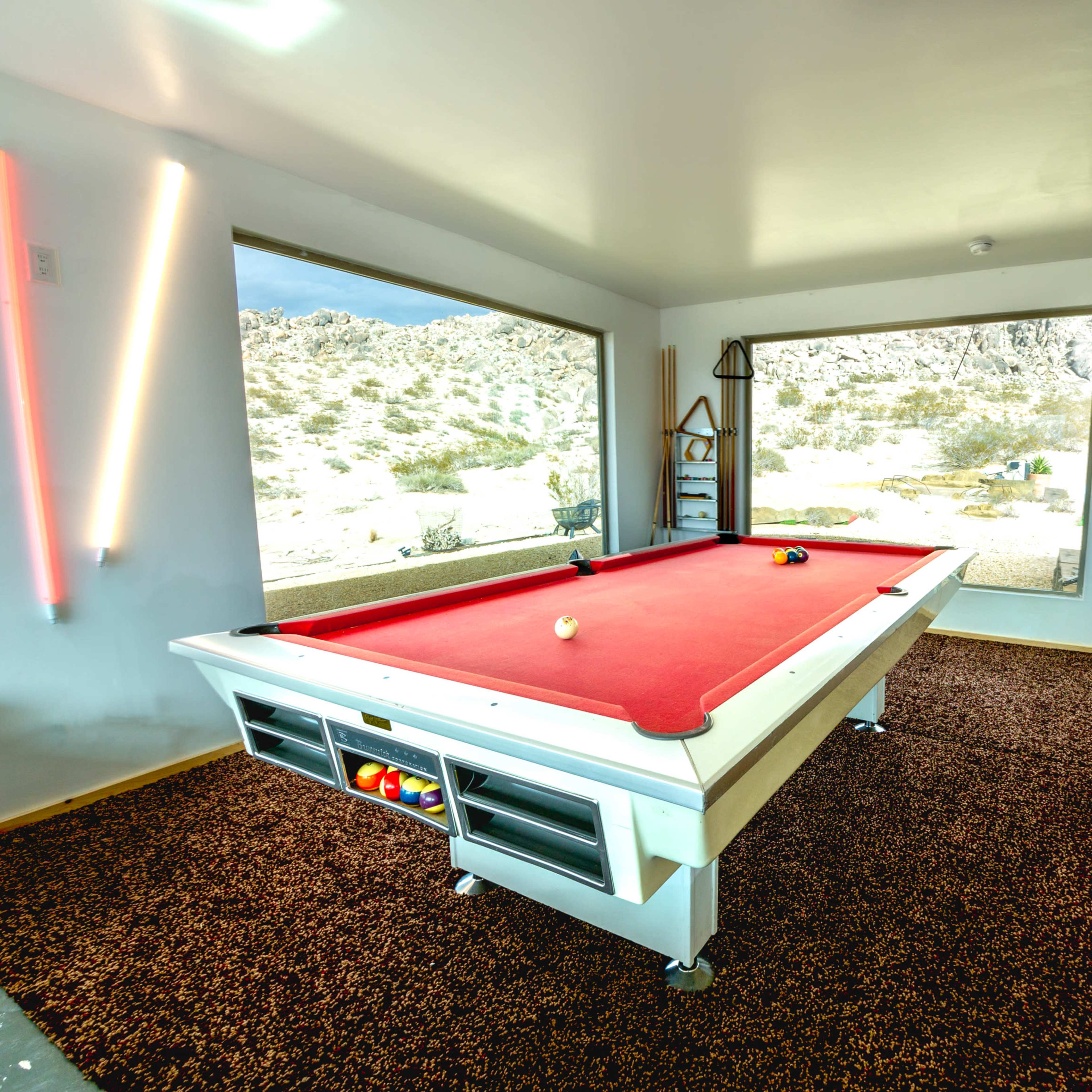 A bright, modern game room features a red pool table positioned between large windows overlooking a rocky landscape.